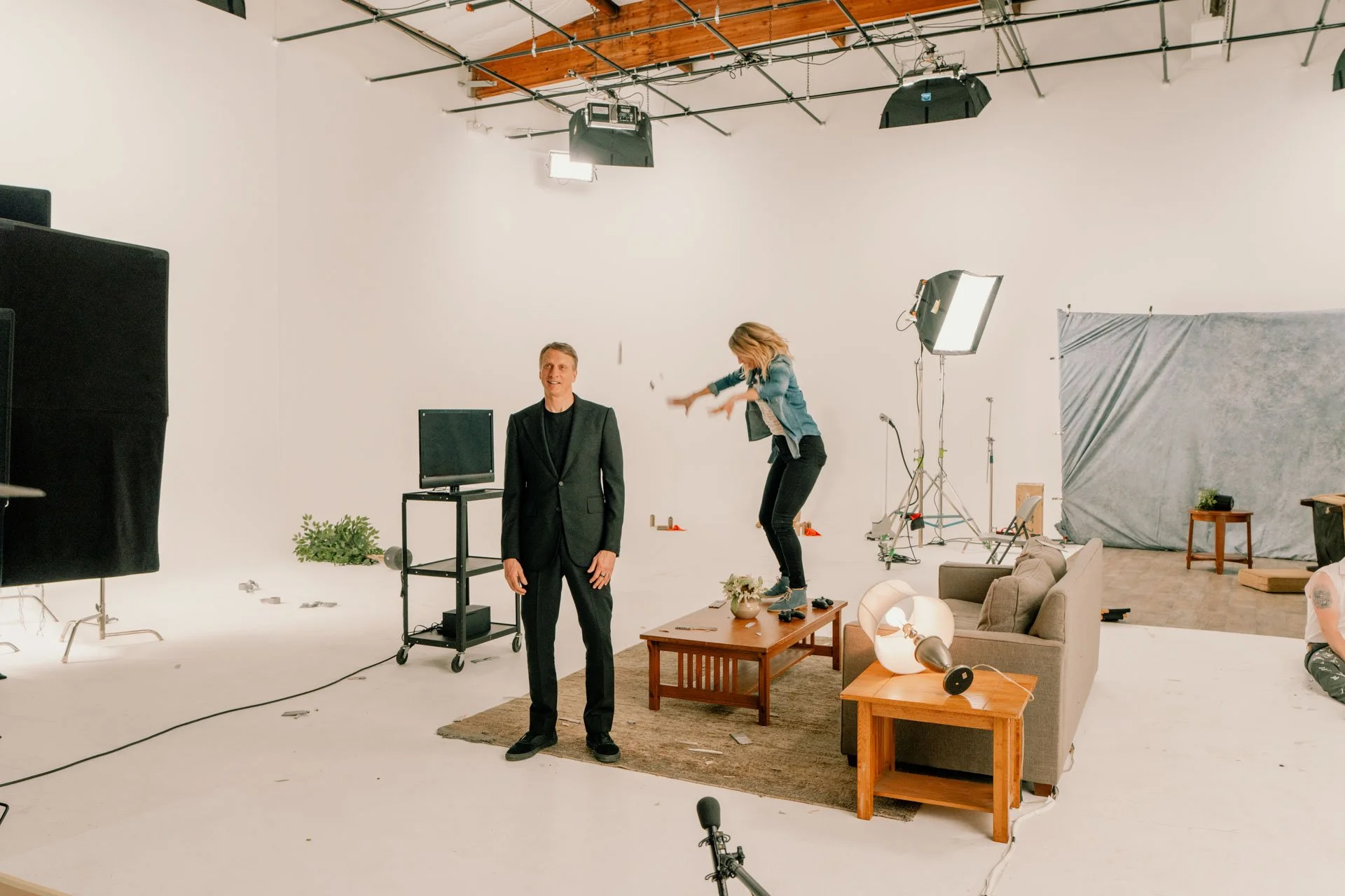 A person standing in front of a green screen while another person jumps off a coffee table in a professional video studio
