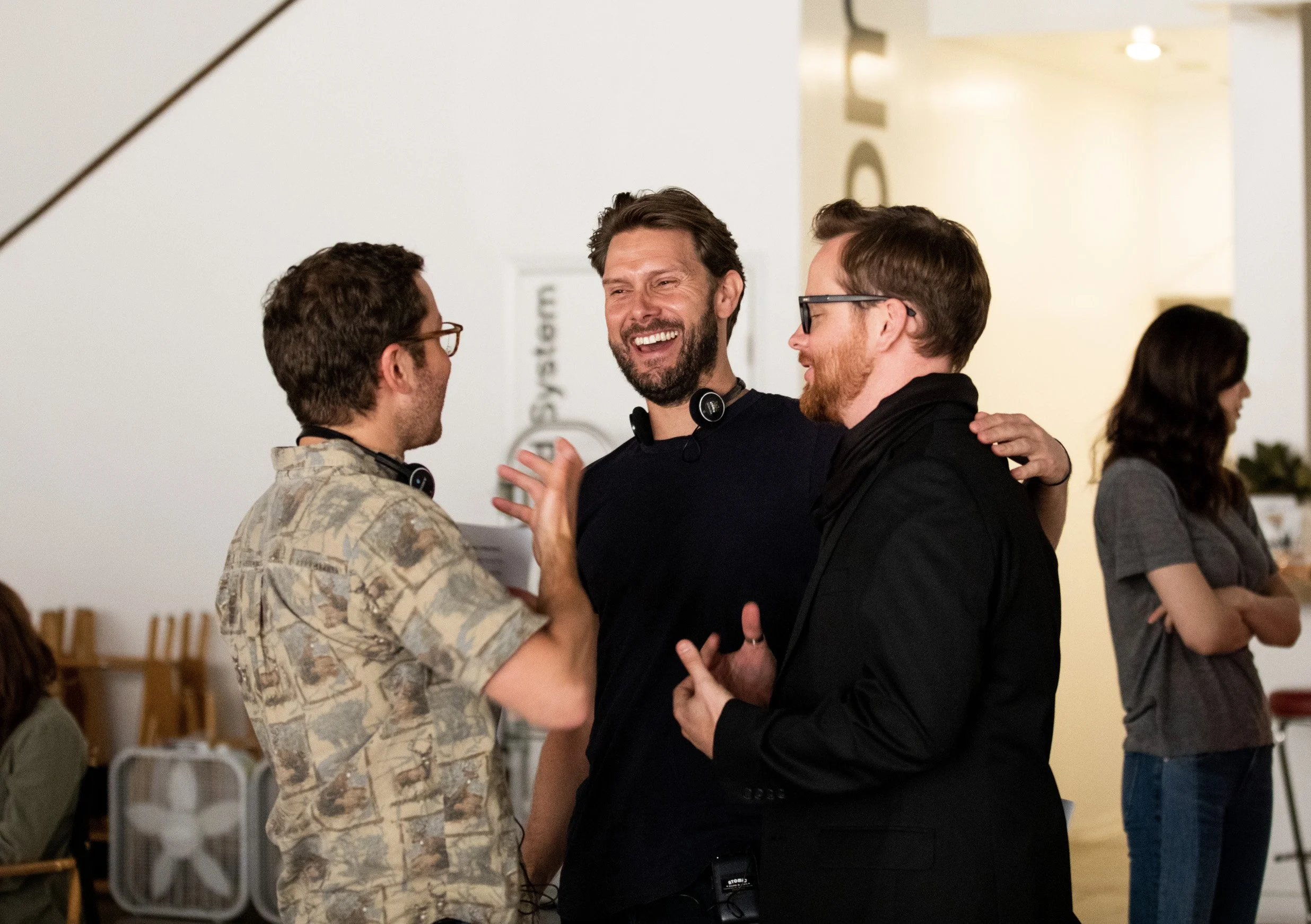 Three men having a lively conversation and smiling, in an indoor setting with others in the background.