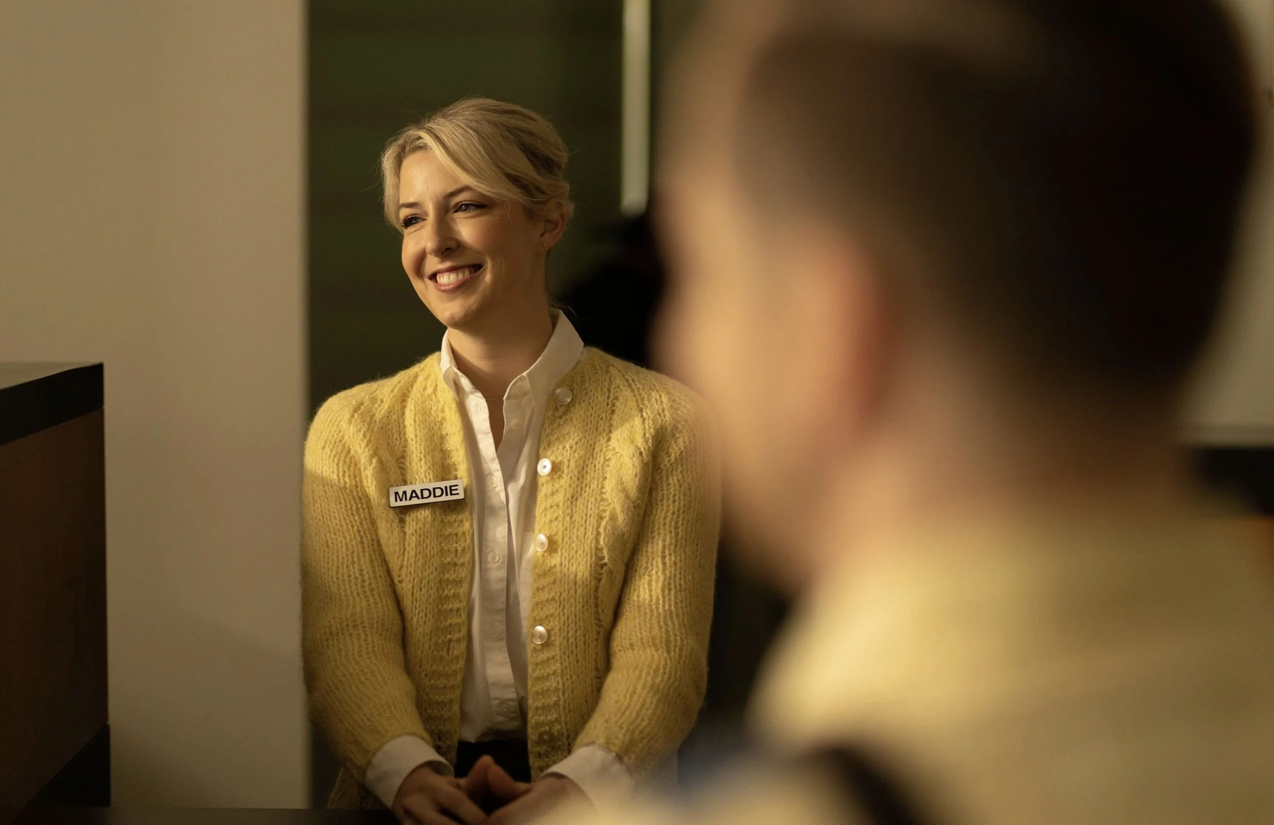 A woman with blonde hair smiling at a man, wearing a yellow sweater with a name badge that reads 'MADDIE'.