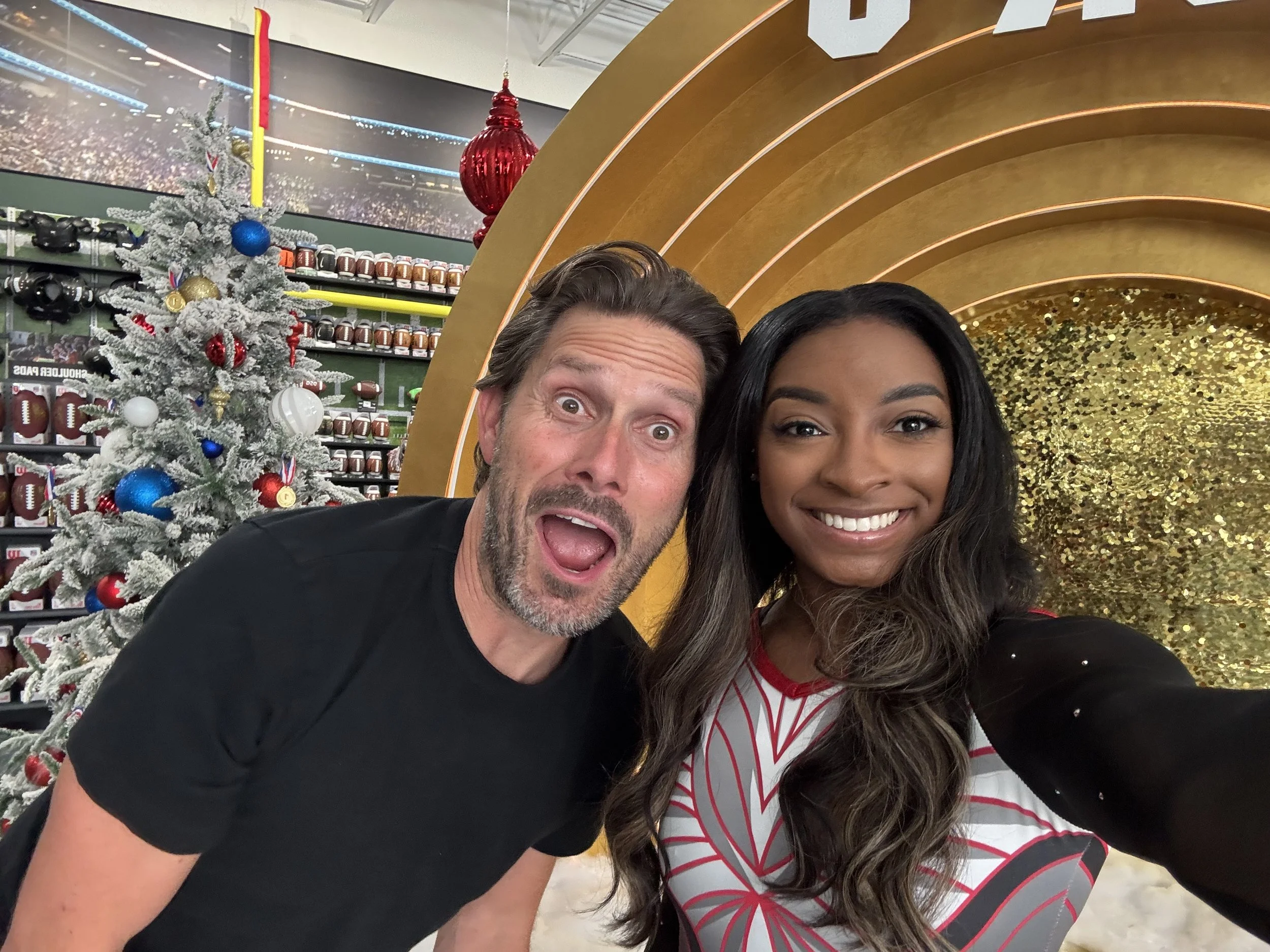 Two people taking a selfie in front of a decorated Christmas tree and gold backdrop, with a sports store and decorations in the background.