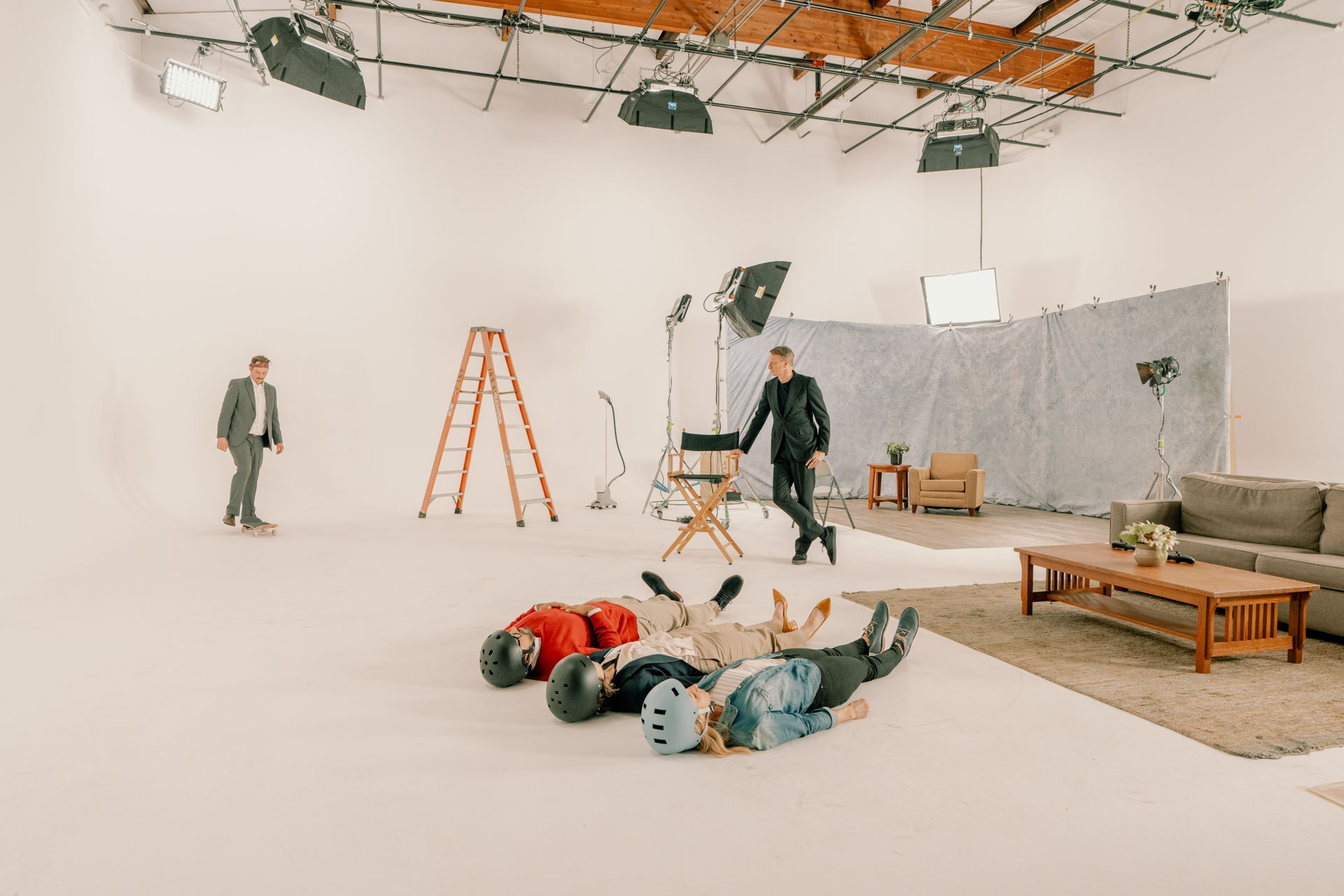 A film or photo shoot setup with three people lying on the floor wearing helmets, a person in a suit standing nearby, and another person in a suit skateboarding in a studio with professional lighting, backdrop, and studio furniture.