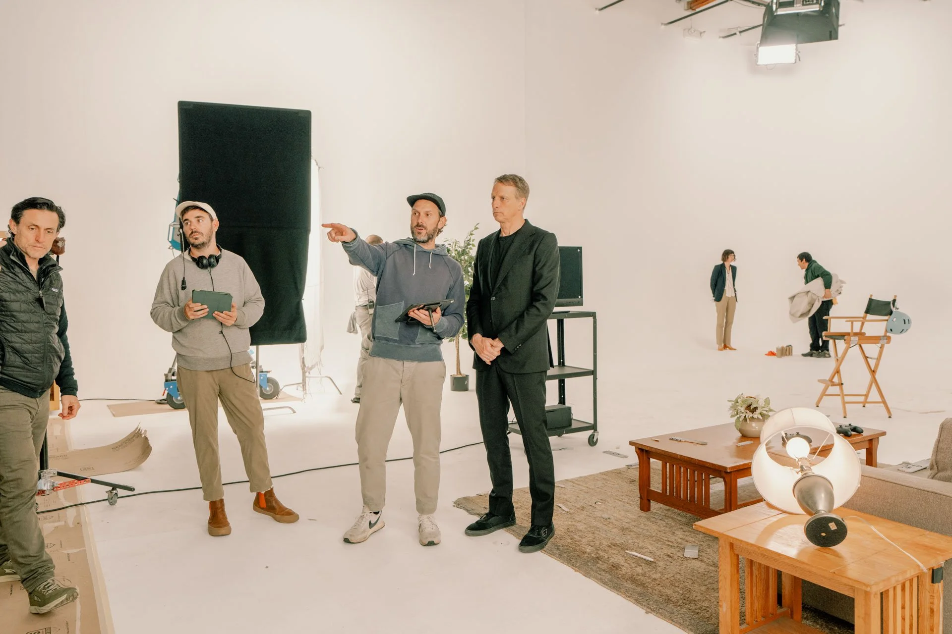 Film crew on set in a white studio with lighting equipment, a large black backdrop, and various props, as actors prepare to shoot a scene.