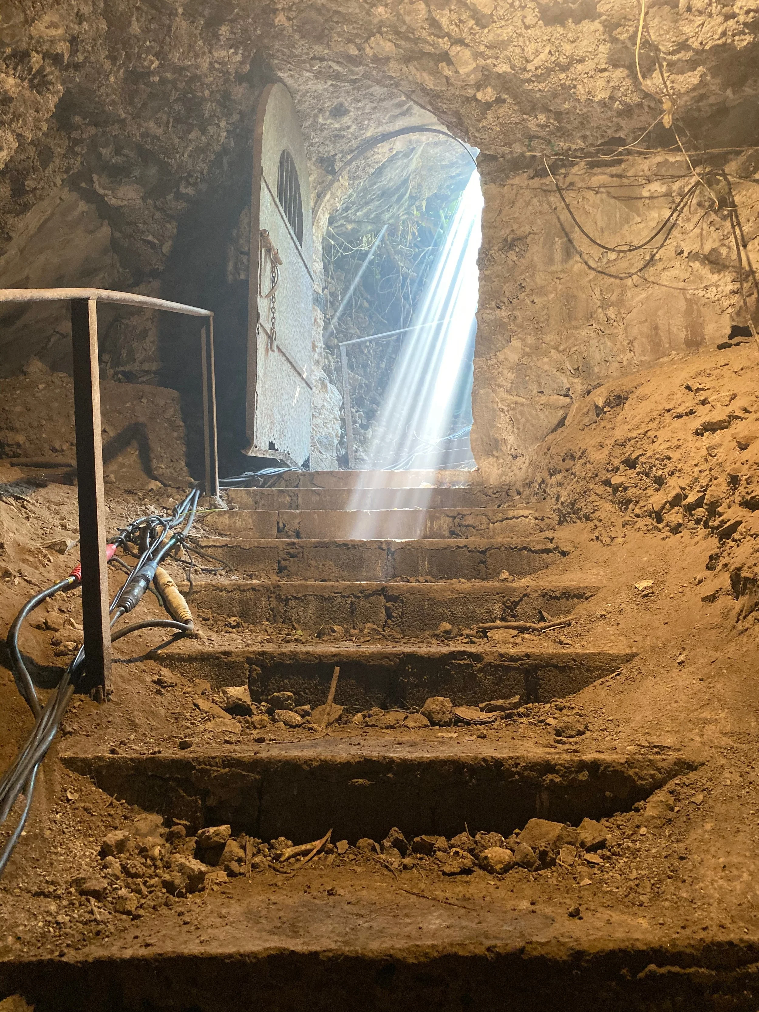 Rough stone staircase leading up to an open doorway with sunlight streaming through, inside a cave or tunnel with dirt walls and exposed wiring.