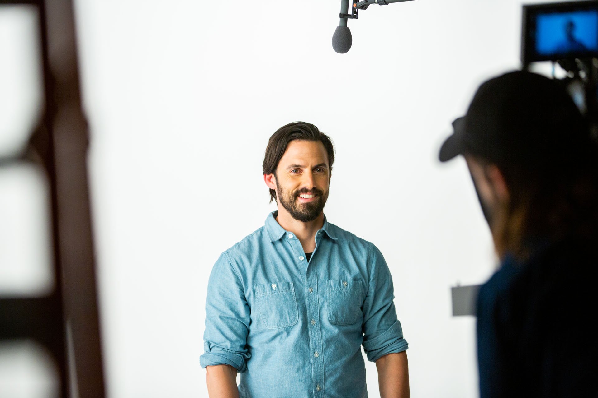 Man with dark hair and beard smiling while standing in front of a white background during filming, with a camera operator and microphone overhead.