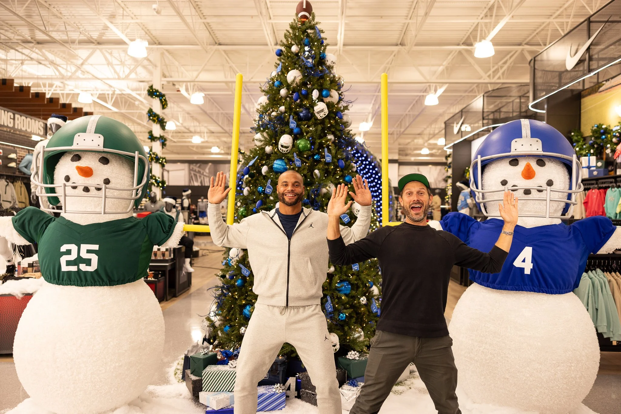 Two men standing in a retail store decorated for Christmas, flanked by two large snowman mascots wearing football helmets and jerseys, with a Christmas tree in the background.