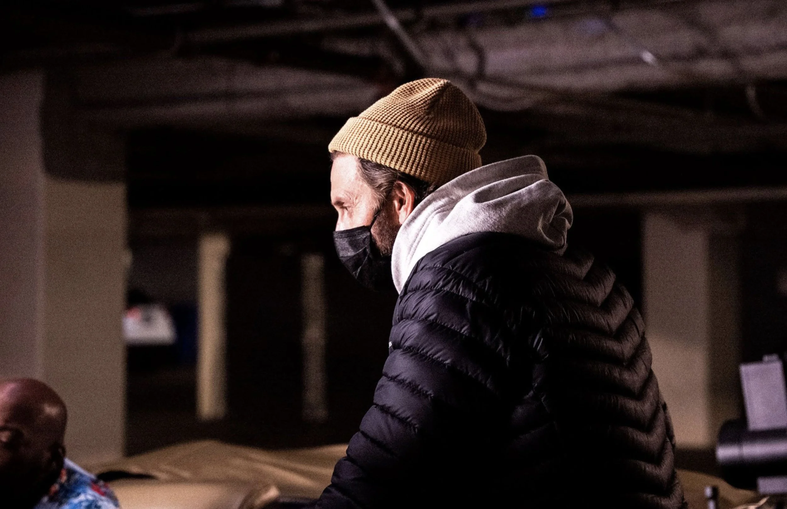A man with a beard wearing a tan knit beanie, a black face mask, a gray hoodie, and a black puffer jacket, standing indoors in a dimly lit room.