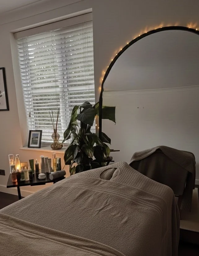 Spa room with massage table covered in beige sheet, large leafy plant, mirror with fairy lights, window with white blinds, and candles on a tray, creating a calming atmosphere.