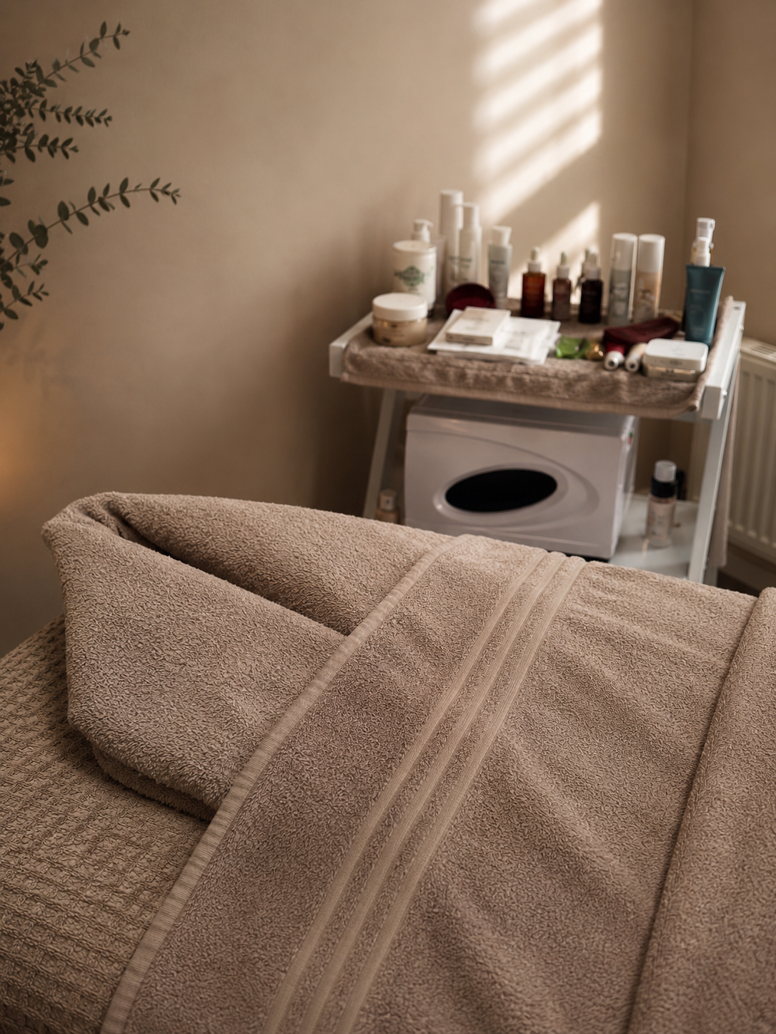 A massage table covered with a beige towel in a spa room. In the background, a white shelf holds skincare and massage products, with sunlight creating shadows on the wall.