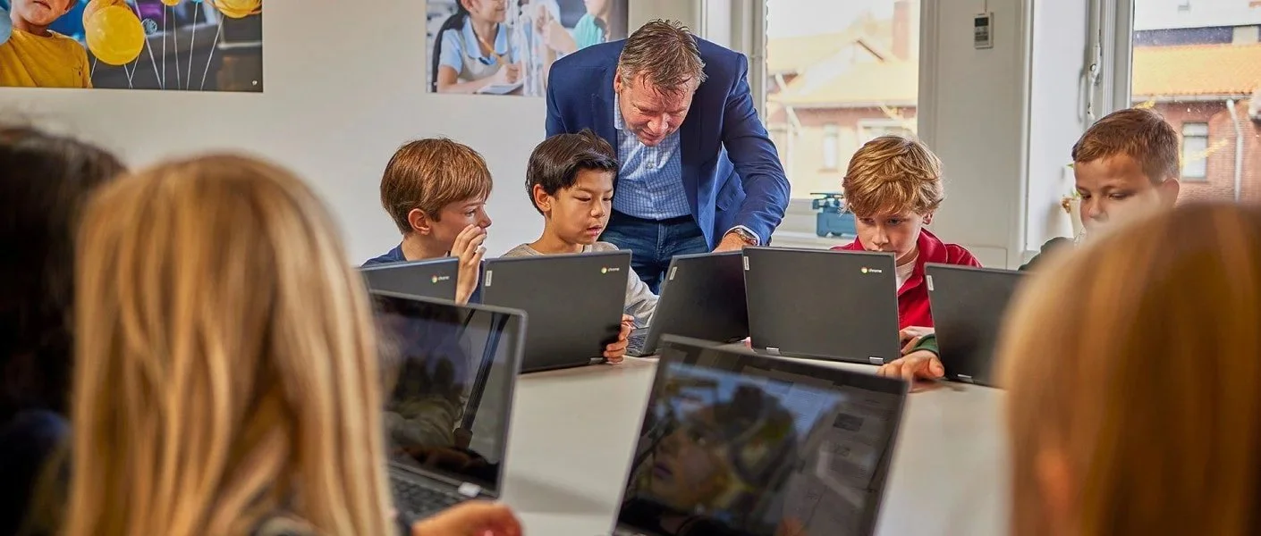 A teacher shows a group of children working on laptops in a classroom with windows and posters on the walls.