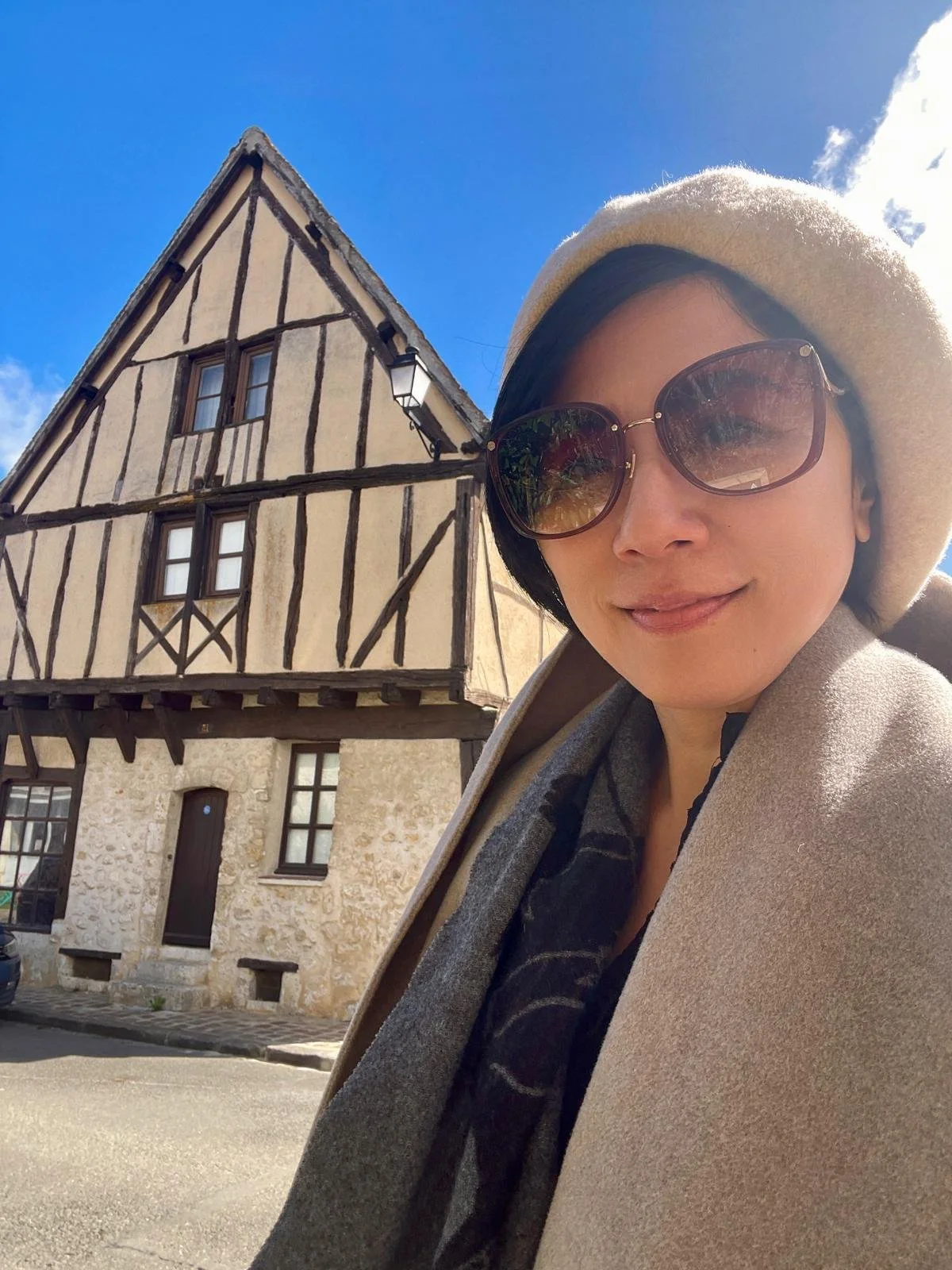 A woman wearing sunglasses and a beige hat taking a selfie in front of a historic half-timbered house under a blue sky with some clouds.