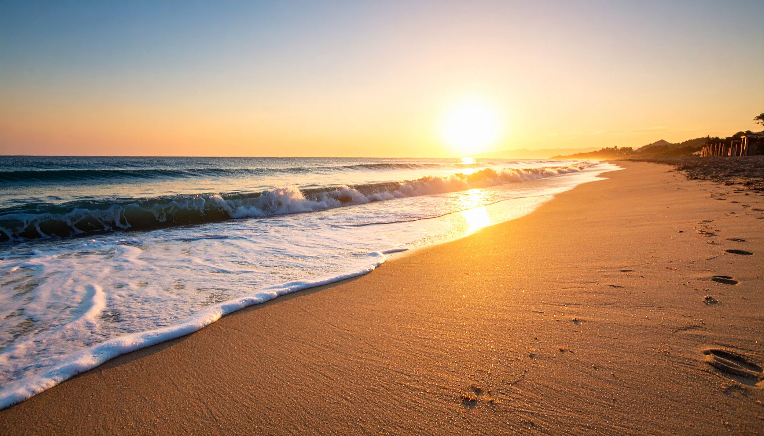 A sandy beach with waves crashing on the shore during sunset, footprints in the sand, and a distant view of hills.