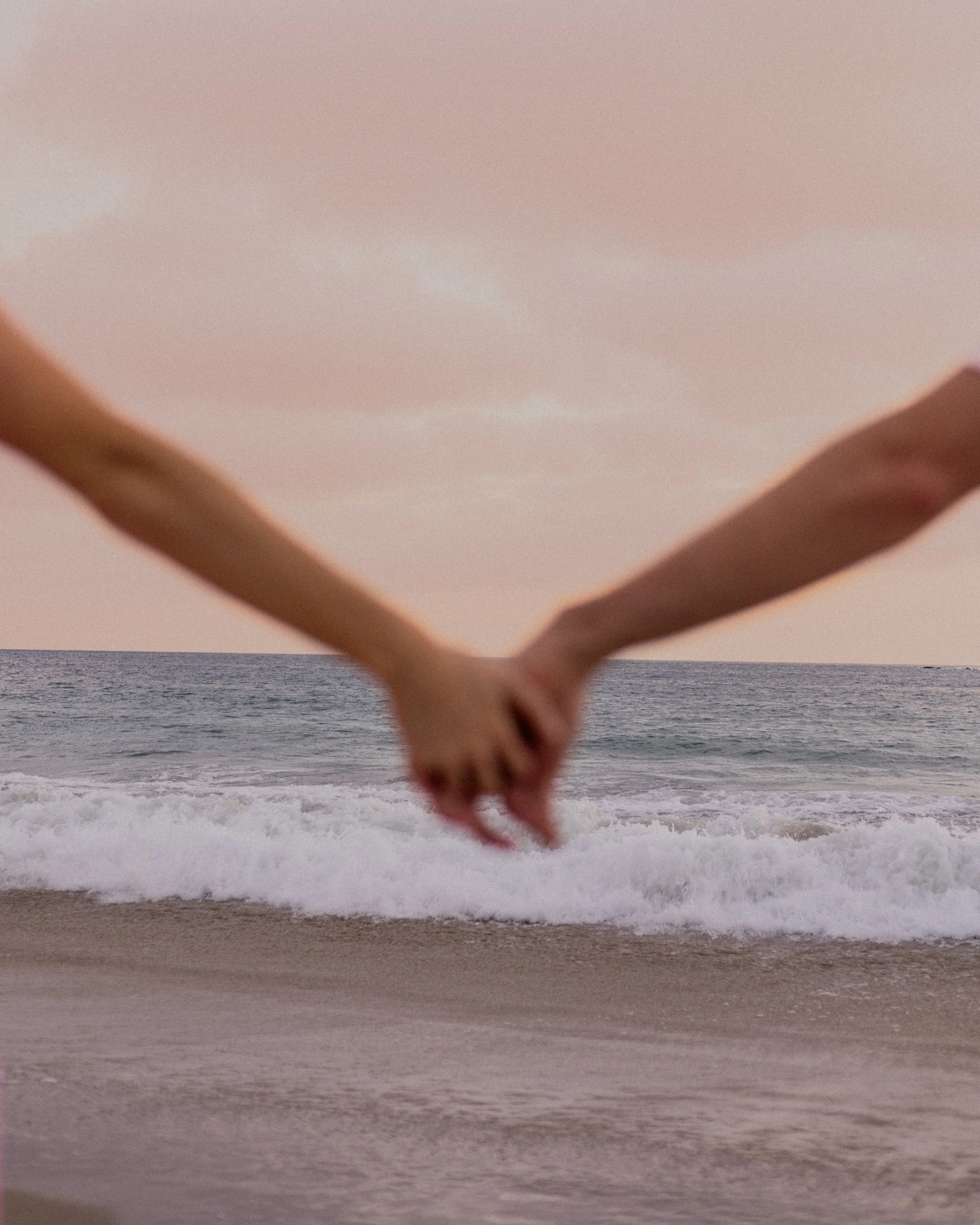 Two people holding hands on the beach at sunset with waves in the background.
