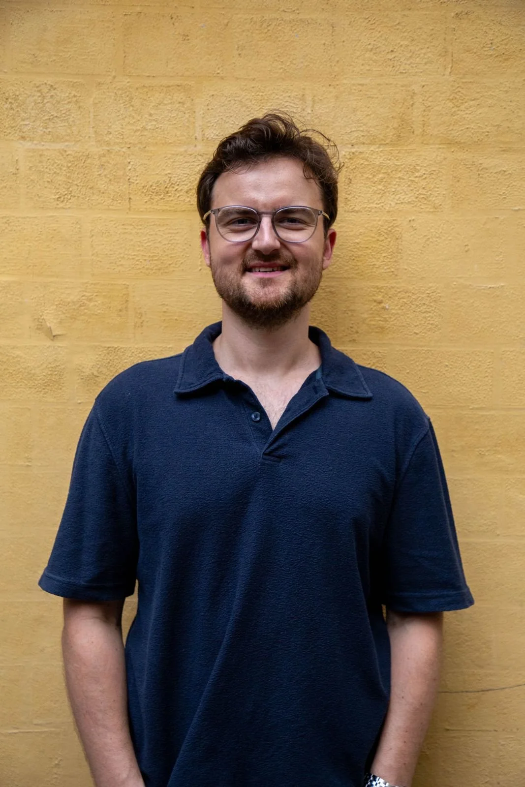 A young man with glasses and a beard smiling, standing in front of a yellow brick wall, wearing a navy blue polo shirt.