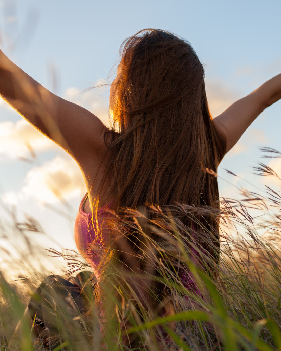 A woman with long brown hair standing in a field of tall grass during sunset, with her arms raised and open to the sky.