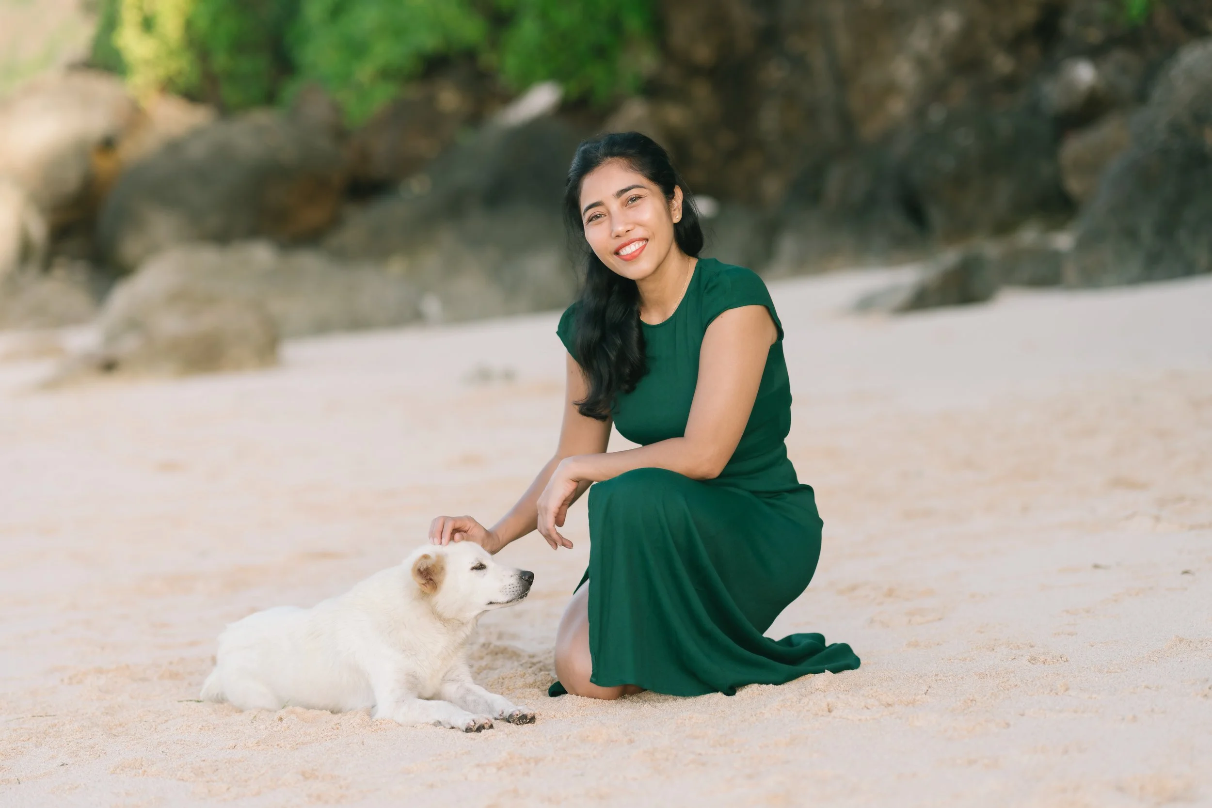 A woman in a green dress kneeling on a sandy beach, petting a white dog lying on the sand.