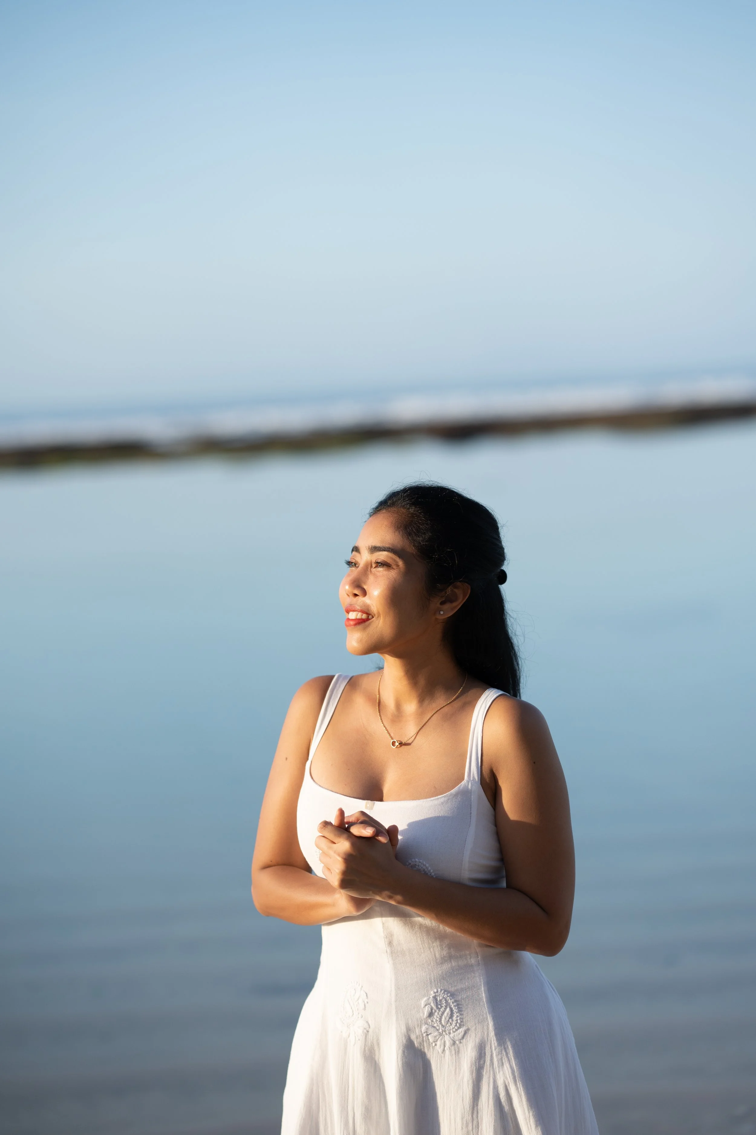 A woman wearing a white dress standing near the water at the beach during daytime, smiling and looking into the distance.