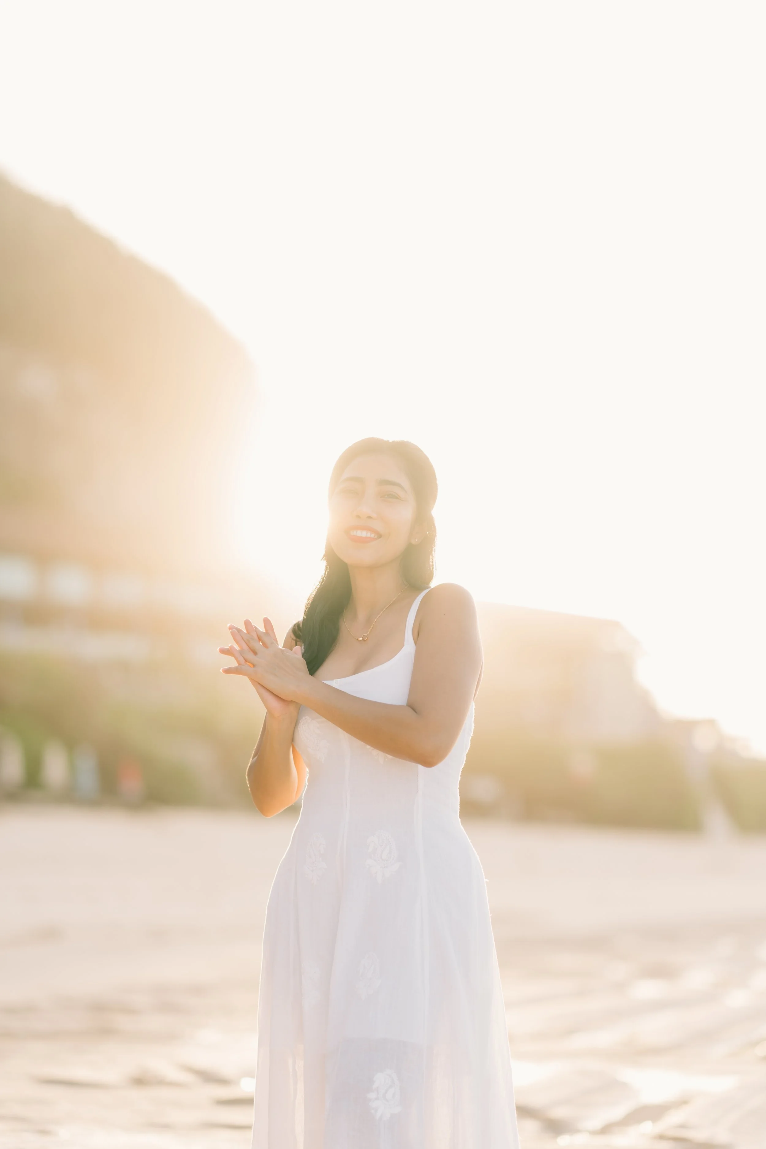 A woman in a white dress standing on a beach at sunset, smiling, with sunlight behind her.