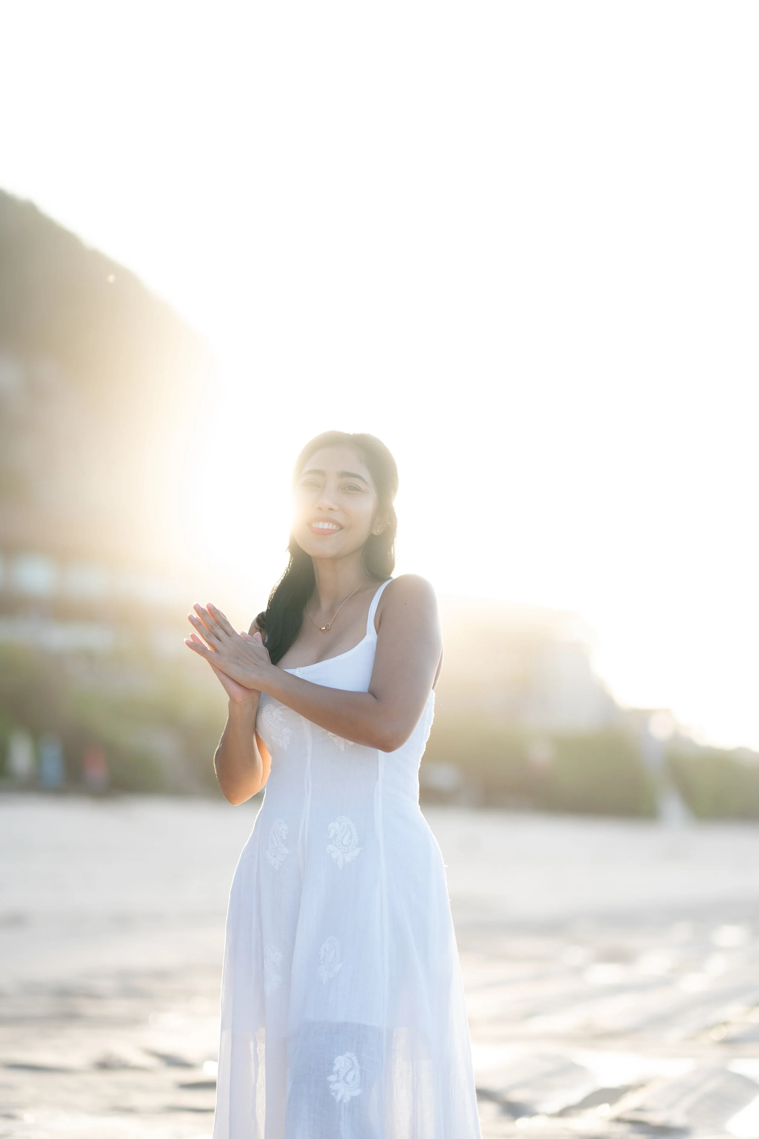 Young woman in a white dress smiling and standing on the beach during sunset.