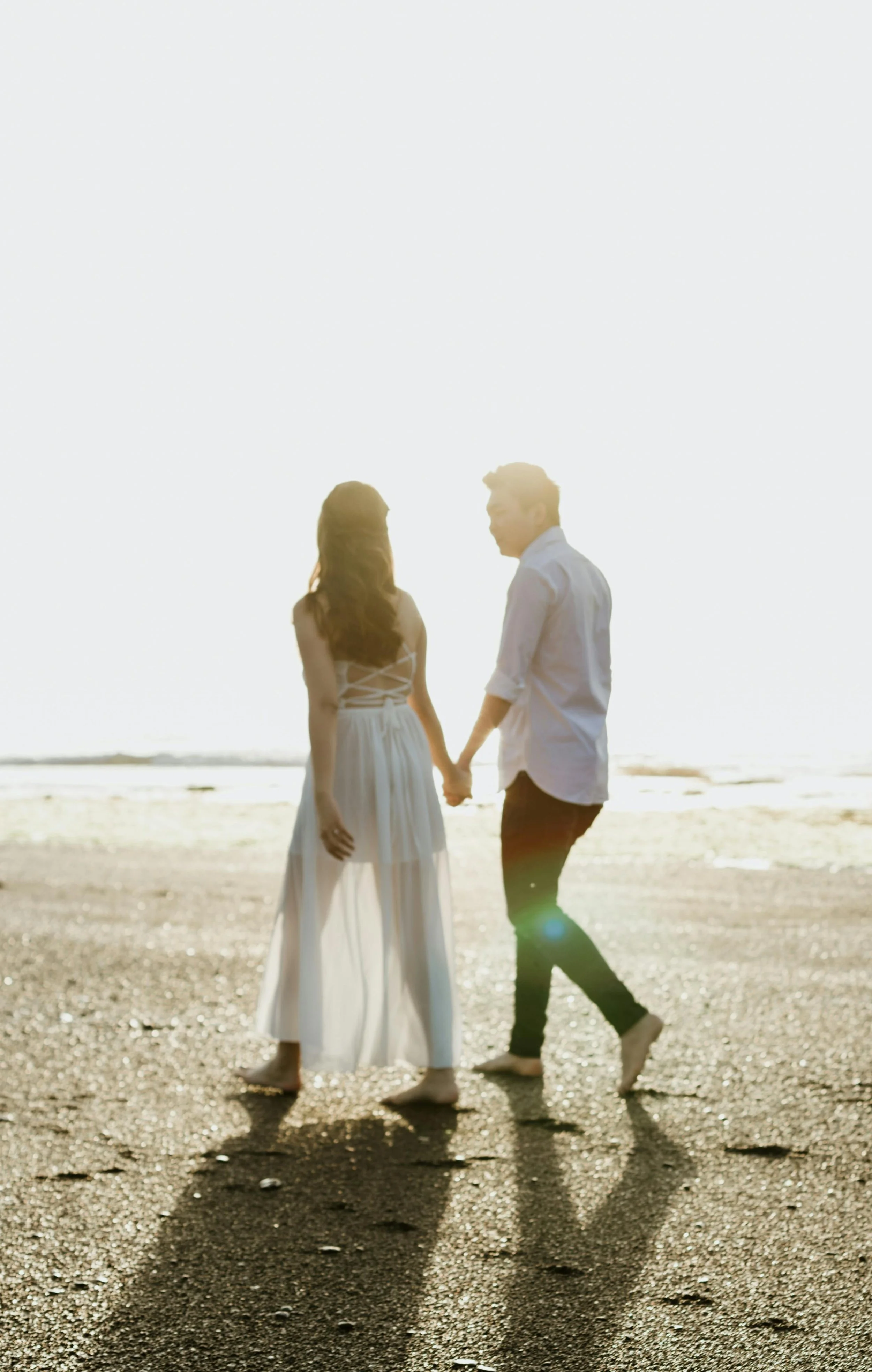 A couple holding hands on a beach during sunset, with their shadows cast on the sand.