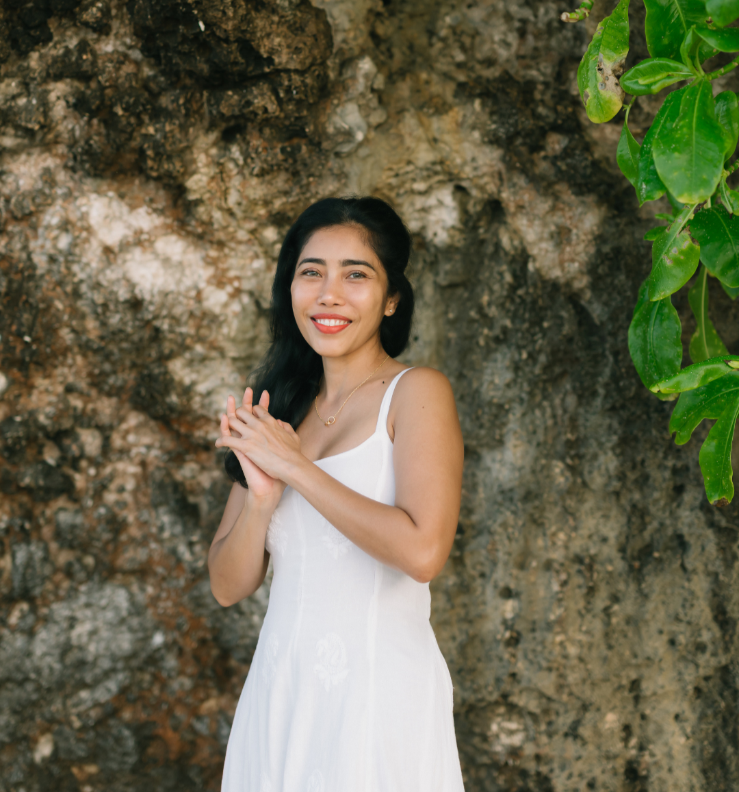 A woman with long black hair in a white dress standing outdoors near a rocky wall, smiling and clasping her hands together.