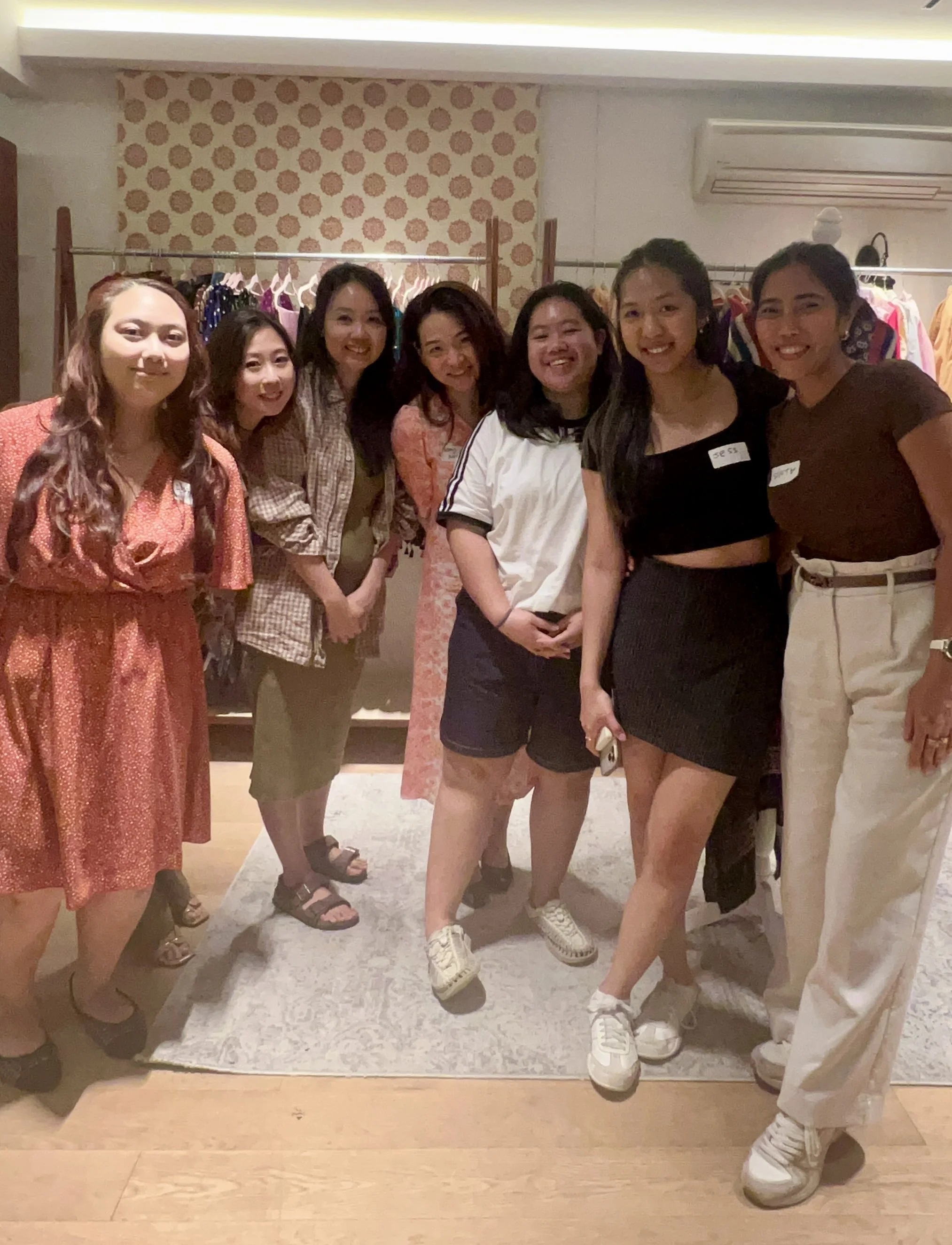 Group of eight women standing indoors, smiling for a photo near a clothing rack and a patterned wall, dressed casually.