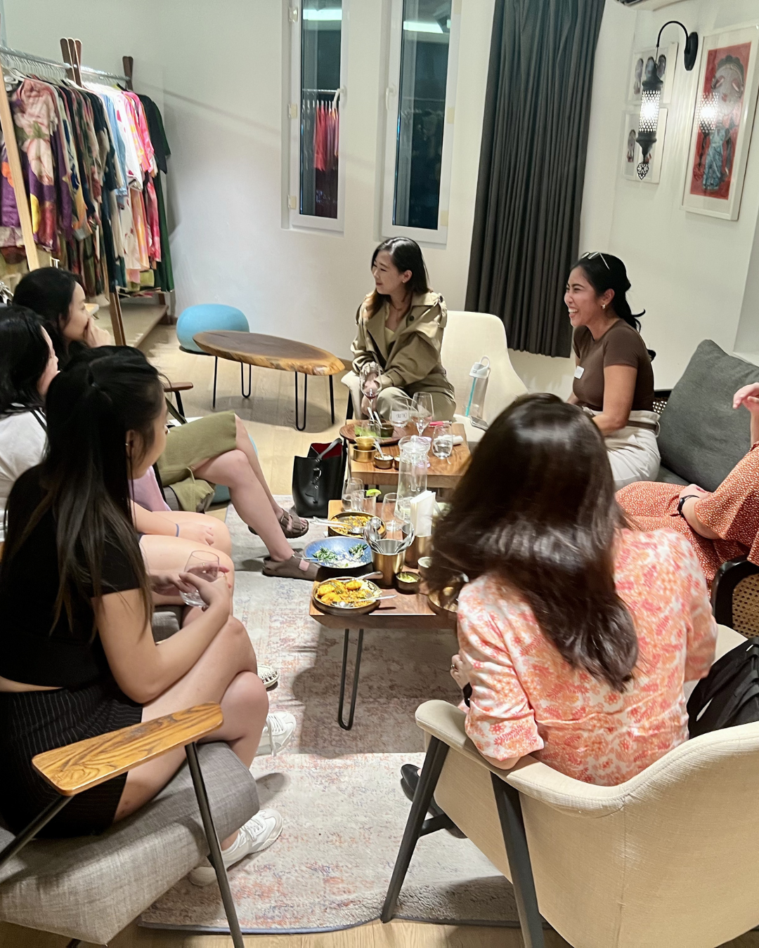 Group of women sitting together in a cozy living room with a rack of colorful clothes in the background, engaging in conversation and enjoying drinks and snacks.