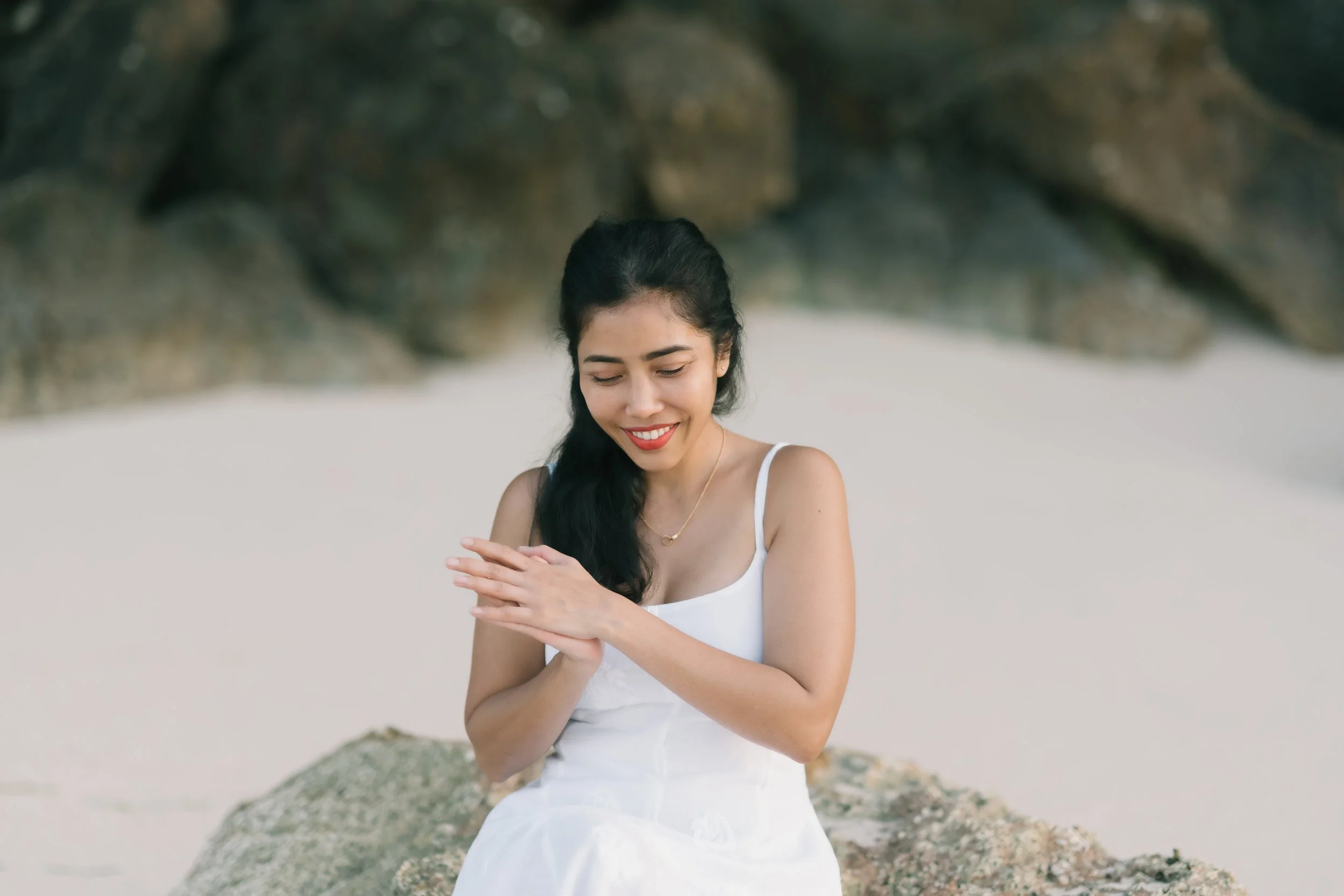 A woman with long dark hair sitting on a rock at the beach, smiling with her eyes closed, wearing a white dress and a gold necklace, with rocks and sand in the background.