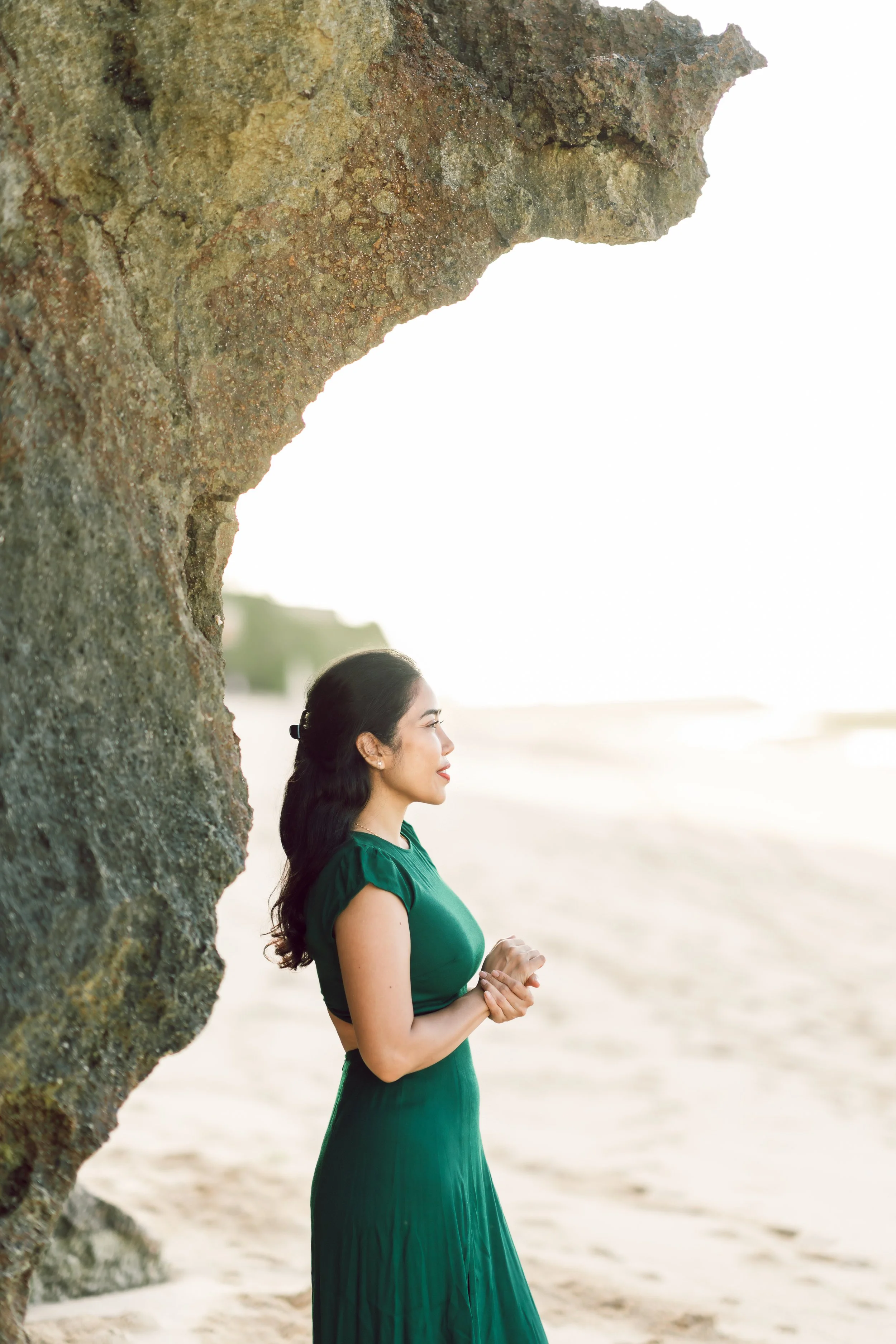 A woman in a green dress standing on a beach near a large rock formation, looking towards the ocean during sunset.