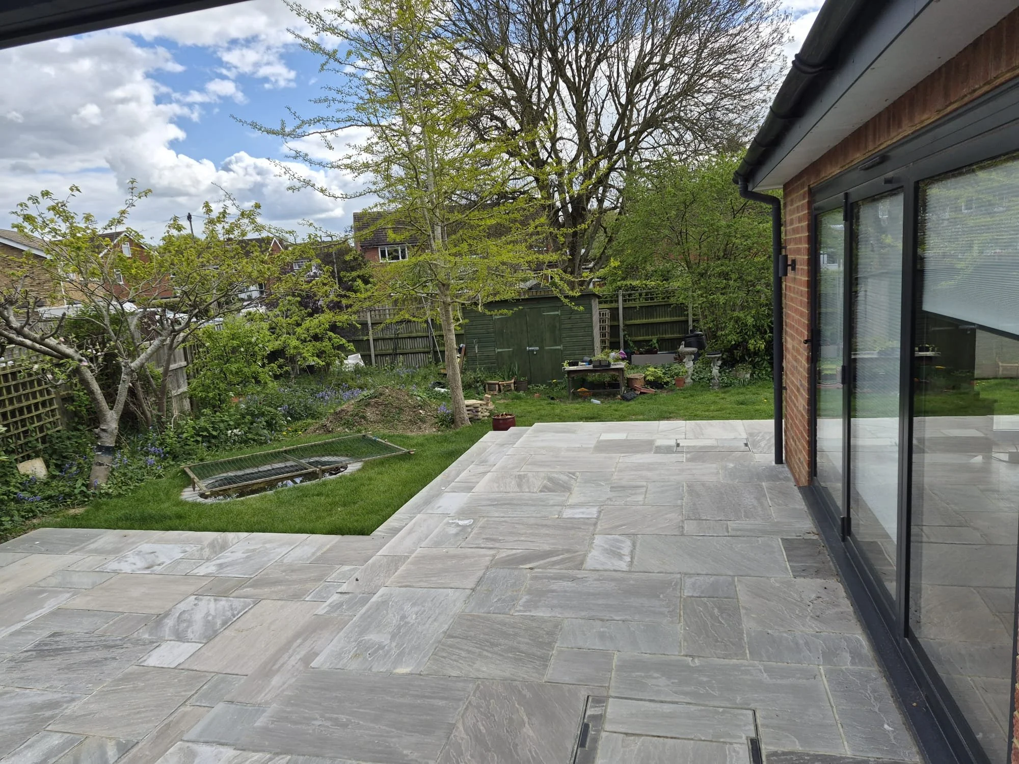 View from inside a house or patio looking out to a backyard with a paved stone patio, grass, trees, and a wooden fence. There is a brick house on the right side with glass doors and a black gutter. The sky is partly cloudy with patches of blue.