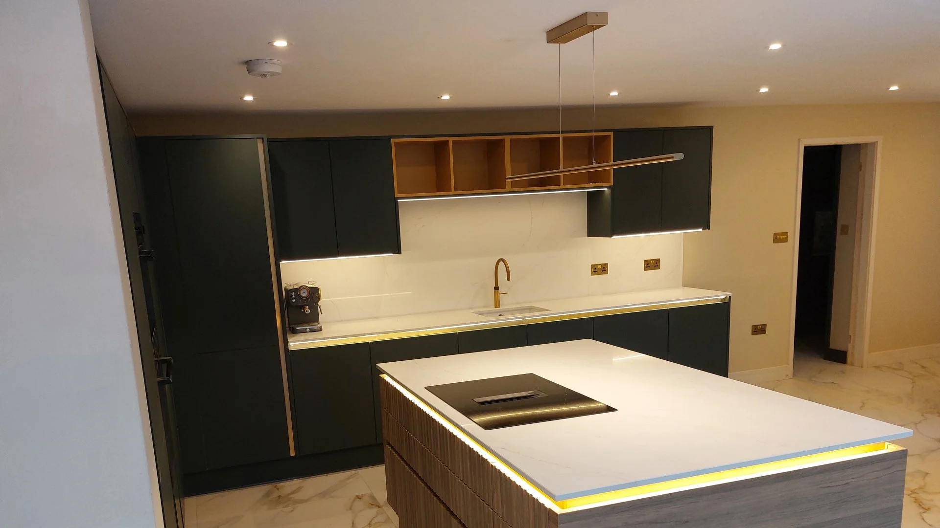 Modern kitchen with black cabinets, white marble countertops, a gold faucet, and a central island with a cooktop. There are open shelves, and built-in lighting accents.