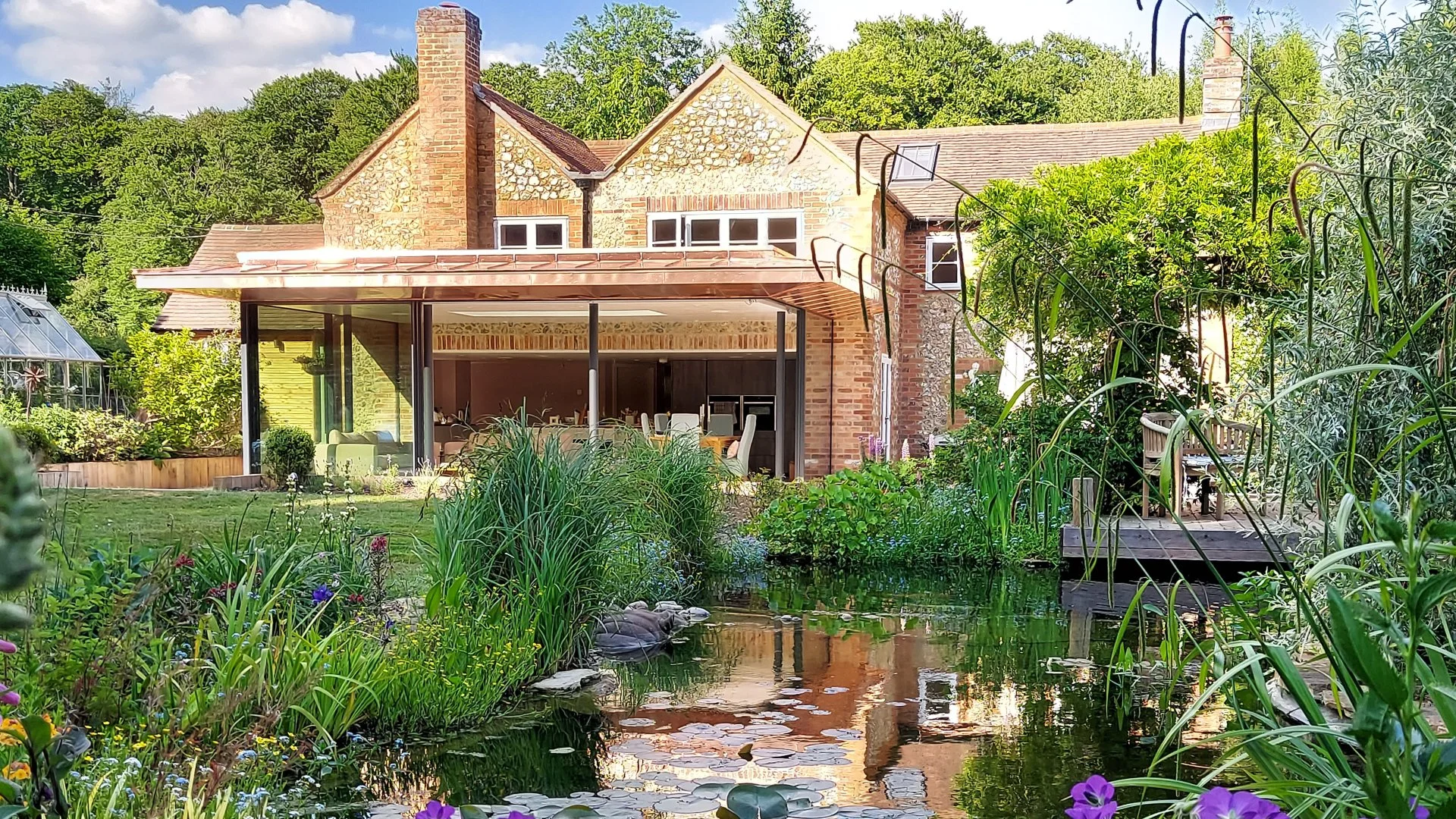 A backyard scene with a pond in the foreground surrounded by various green plants and flowers, a wooden deck on the right, and a house with brick and stone exterior in the background amid tall trees.