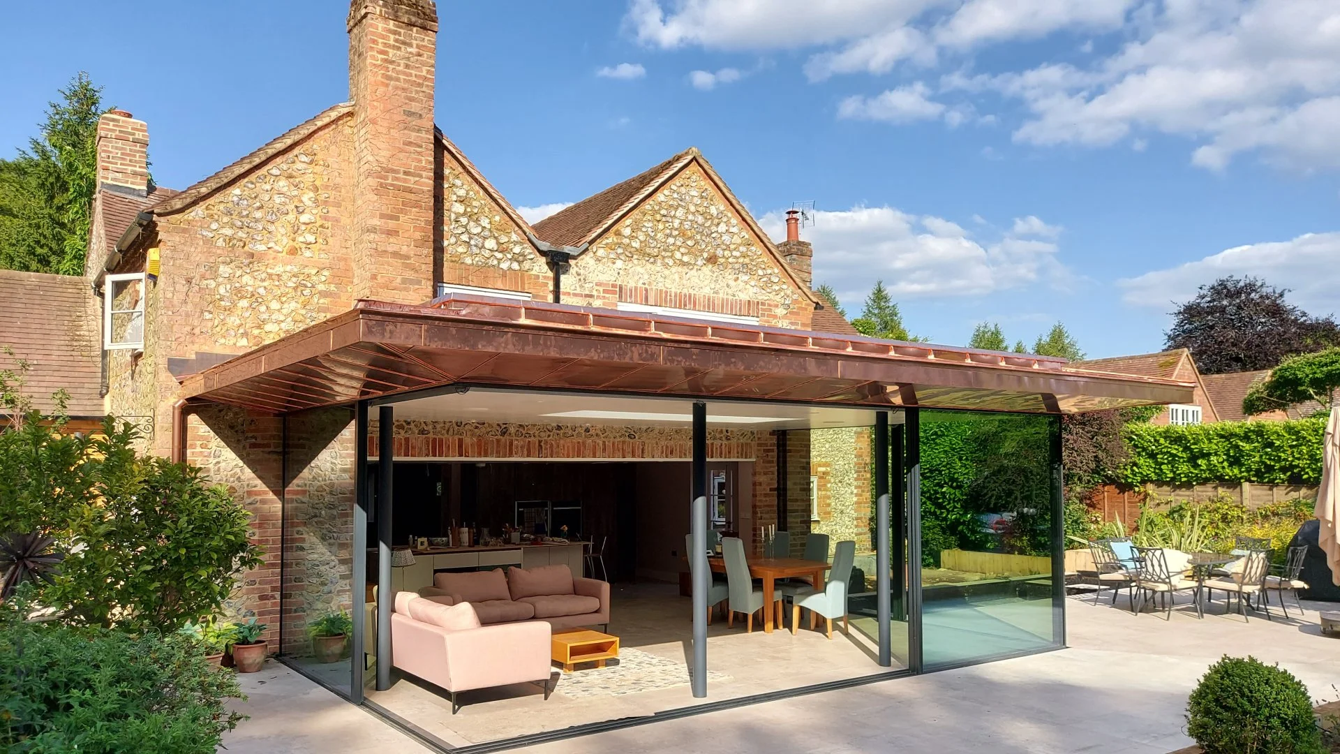 A backyard patio area featuring a modern glass enclosure attached to a brick house with stone accents. The patio has outdoor furniture including a pink couch, dining table with chairs, and some lounge chairs. Green bushes and trees surround the area,