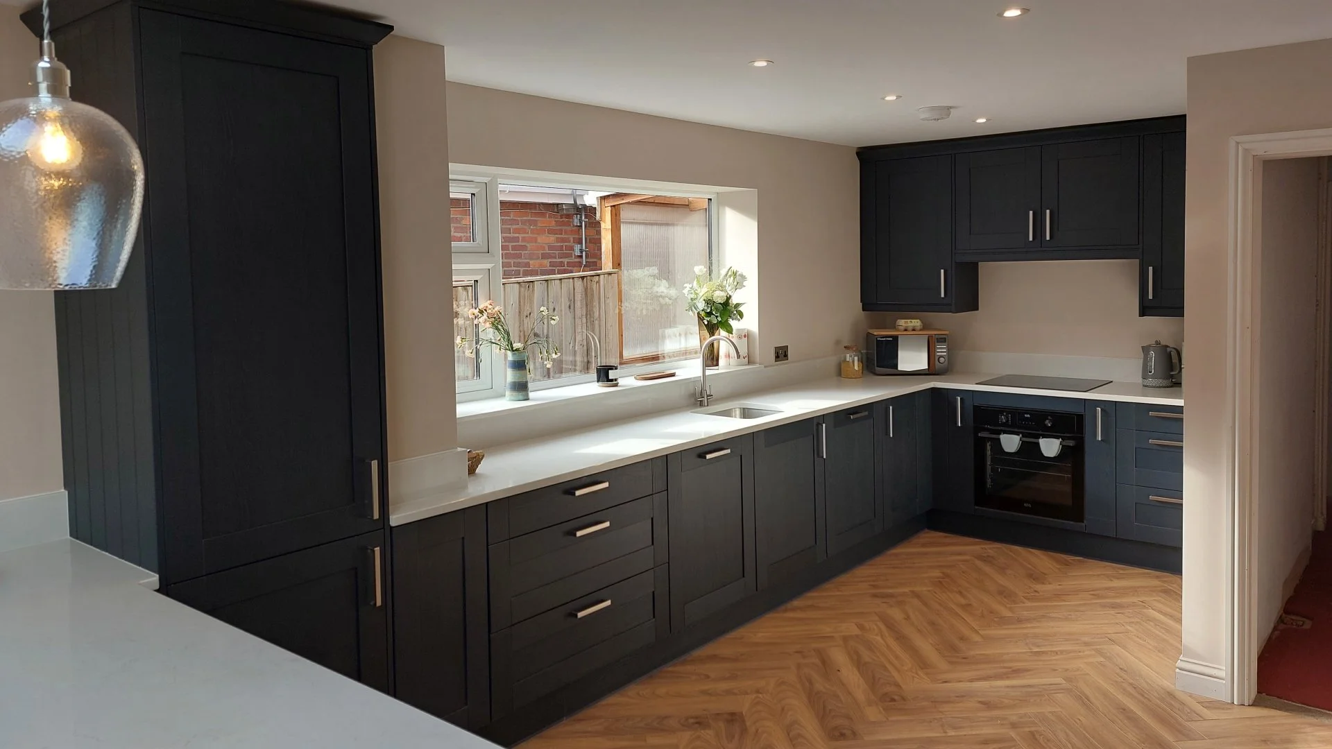 Modern kitchen with black cabinets, white countertops, and a large window with flowers on the sill. Appliances include a microwave, oven, and kettle. The floor is wooden and the ceiling has recessed lighting.