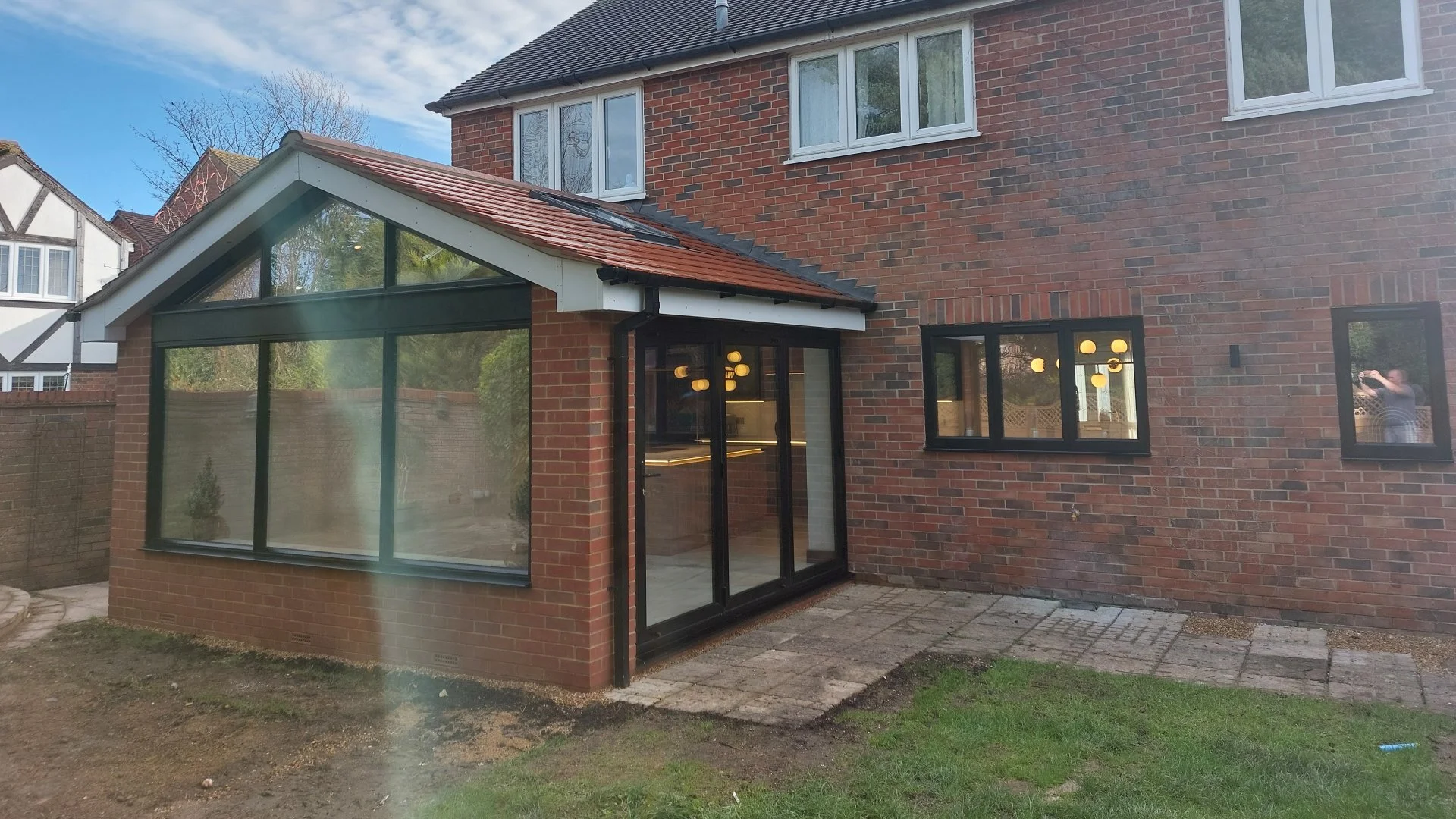Rear view of a brick house with a conservatory extension featuring large glass windows and sliding doors. The house has a pitched roof with tiles, and neighboring houses are visible in the background. Reflections of the person taking the photo are se