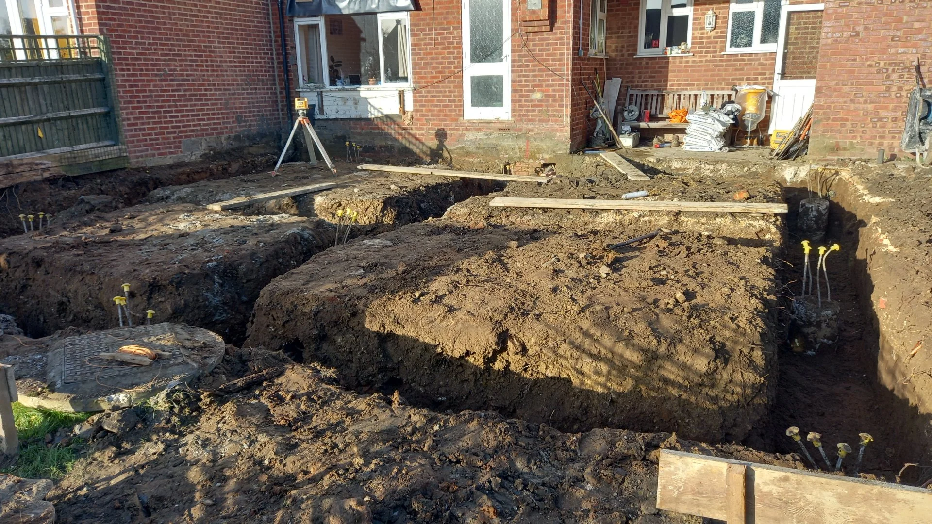 Excavation site in the backyard of a brick house, with trenches, construction materials, and tools for a building project.