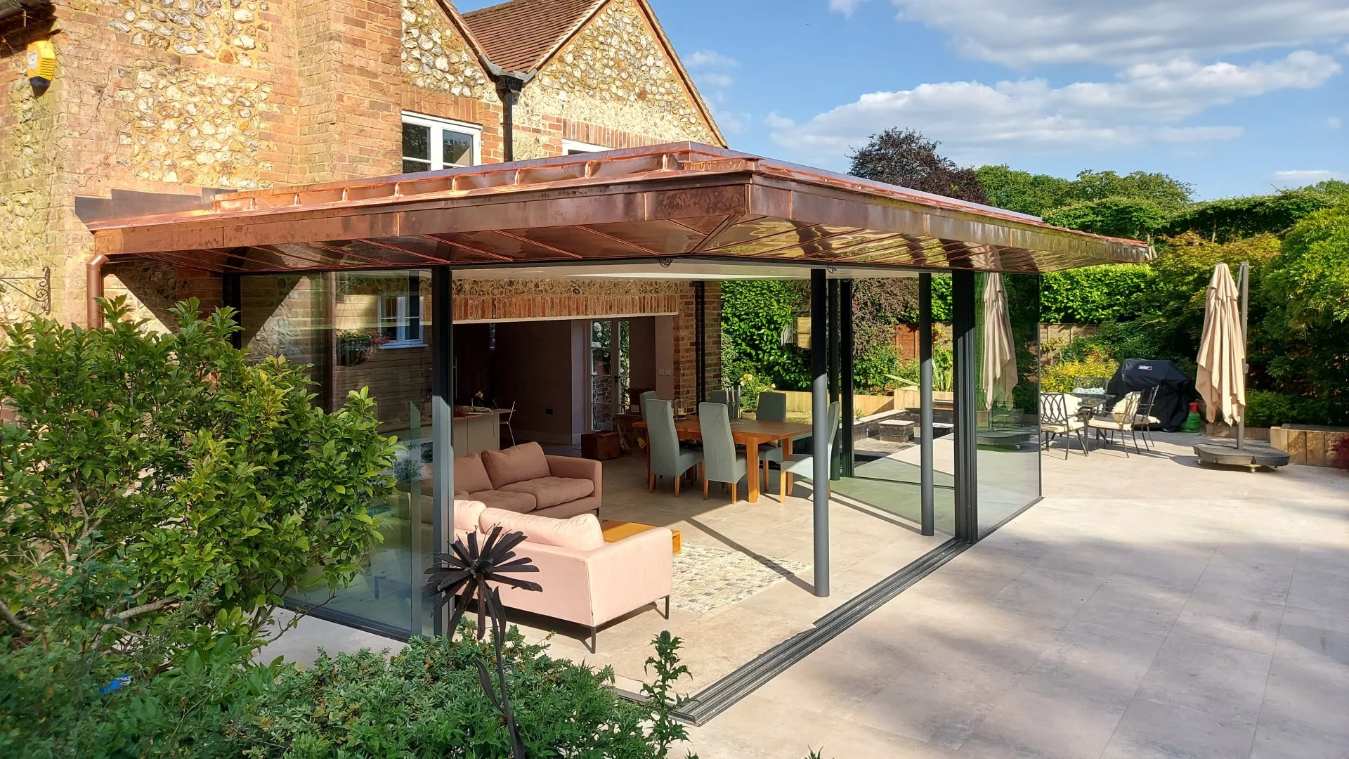 A backyard with a glass enclosure attached to a brick house with a copper roof. Inside is outdoor furniture including a pink sofa and a dining table with chairs. Outside are umbrellas, a barbecue grill, and patio furniture with lush greenery and tree