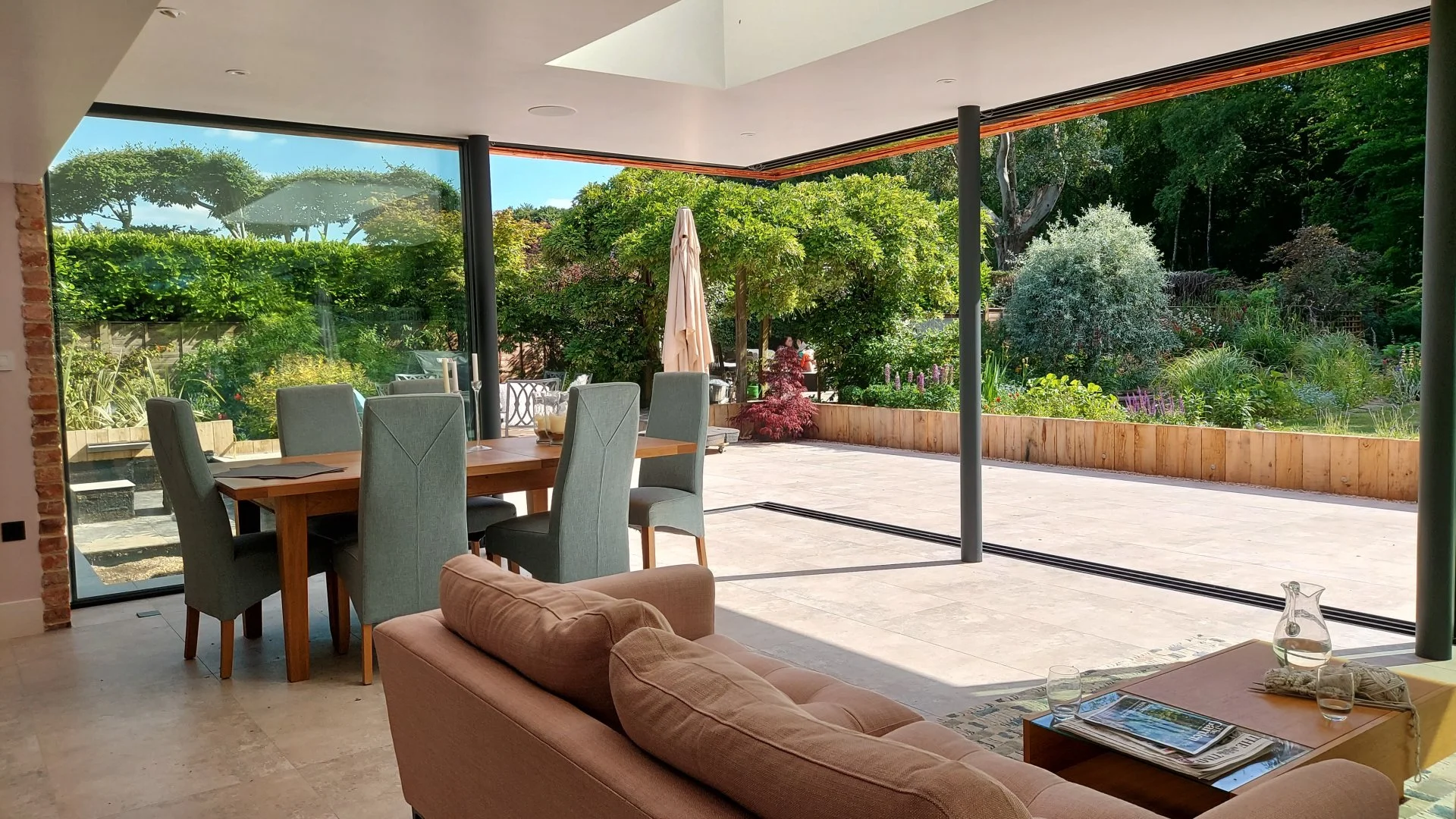 Living room with large glass sliding doors opening to a garden with lush trees and flowering bushes. Indoors, there's a wooden dining table with six gray chairs, a beige sofa with cushions, a small table with magazines, and a glass pitcher with glass