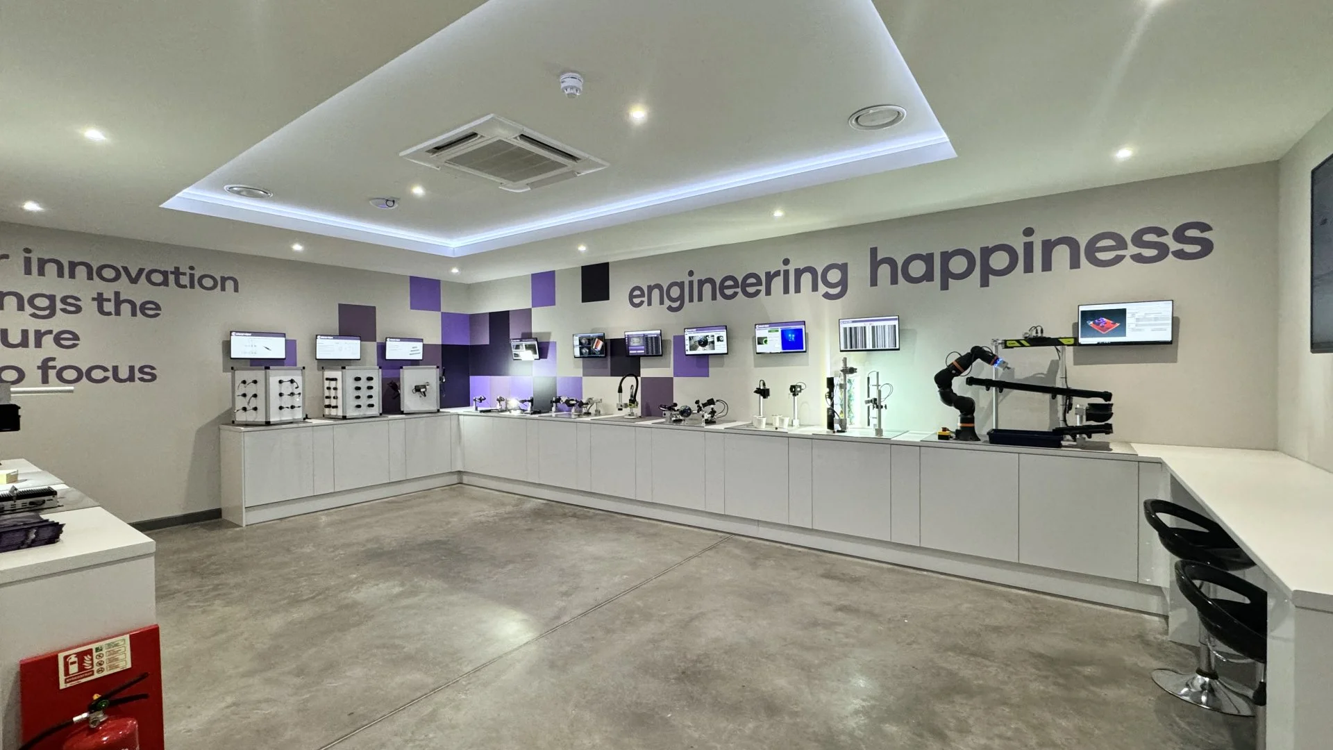 A room with white display counters showcasing various engineering and robotic devices under the slogan 'engineering happiness.' The wall features purple and black geometric patterns and text about innovation and focus.