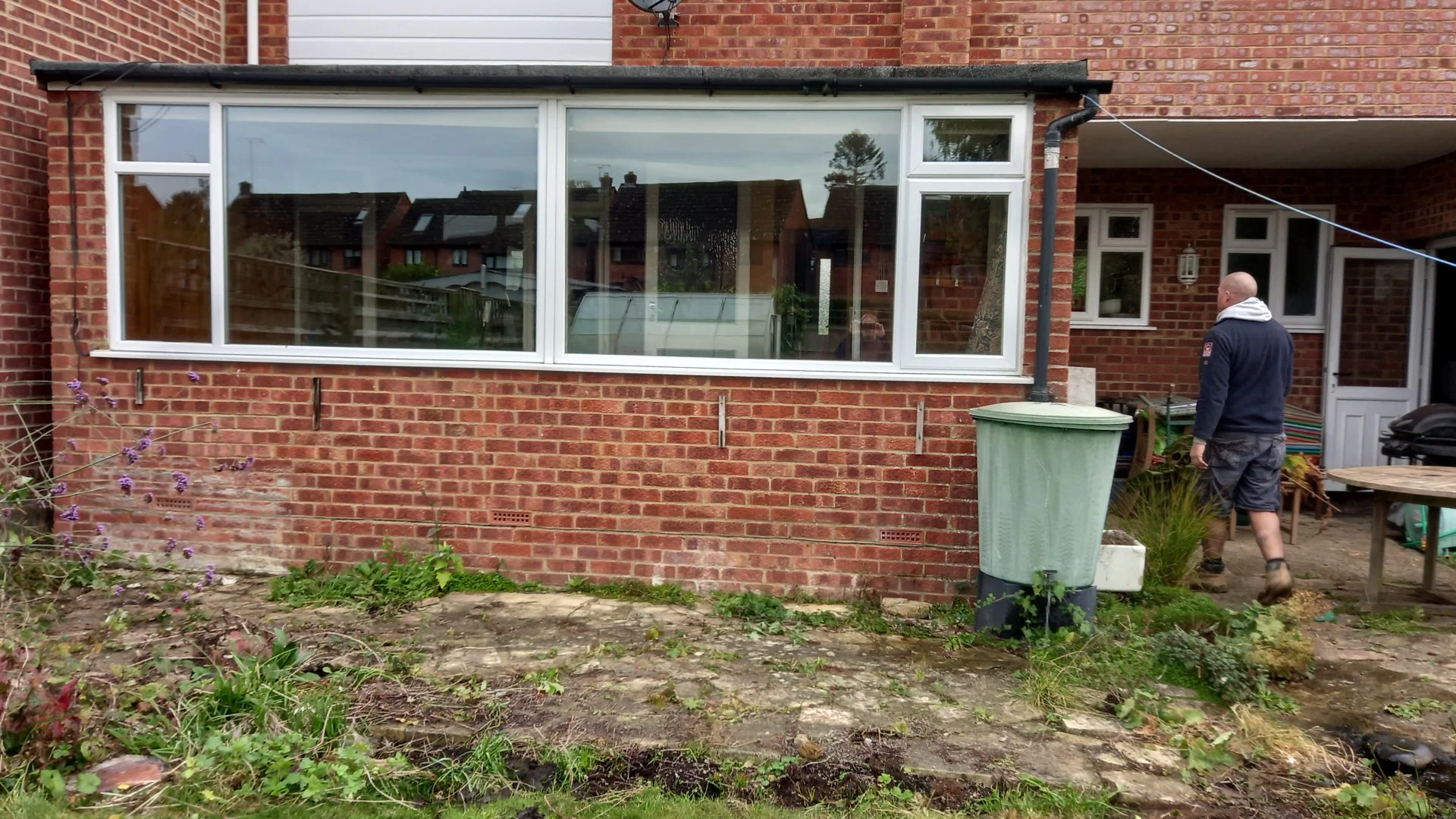 Backyard with red brick house, large window, person with bald head and hoodie, garden, plants, and green trash bin.