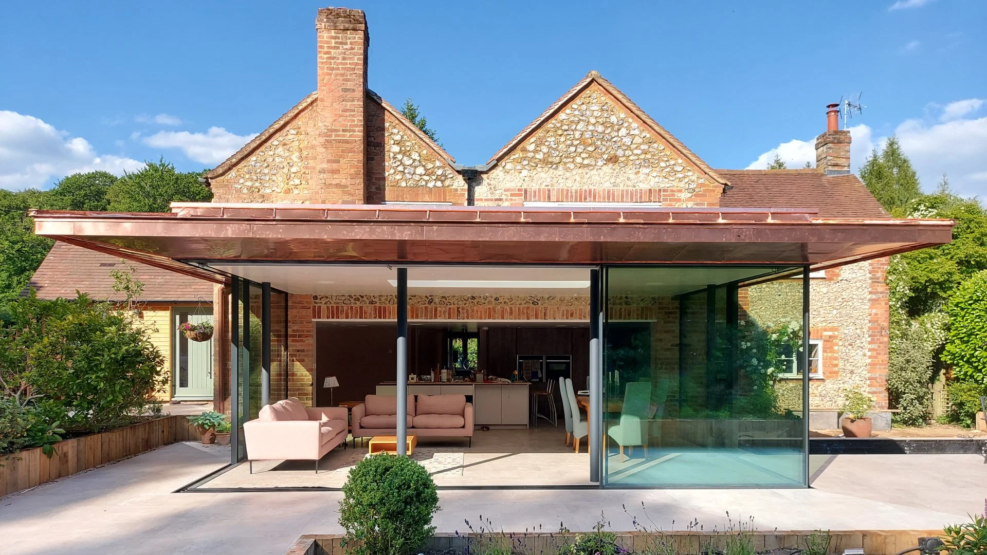 Modern glass extension on brick house with outdoor living space, including pink sofas, a small table, and surrounding plants.