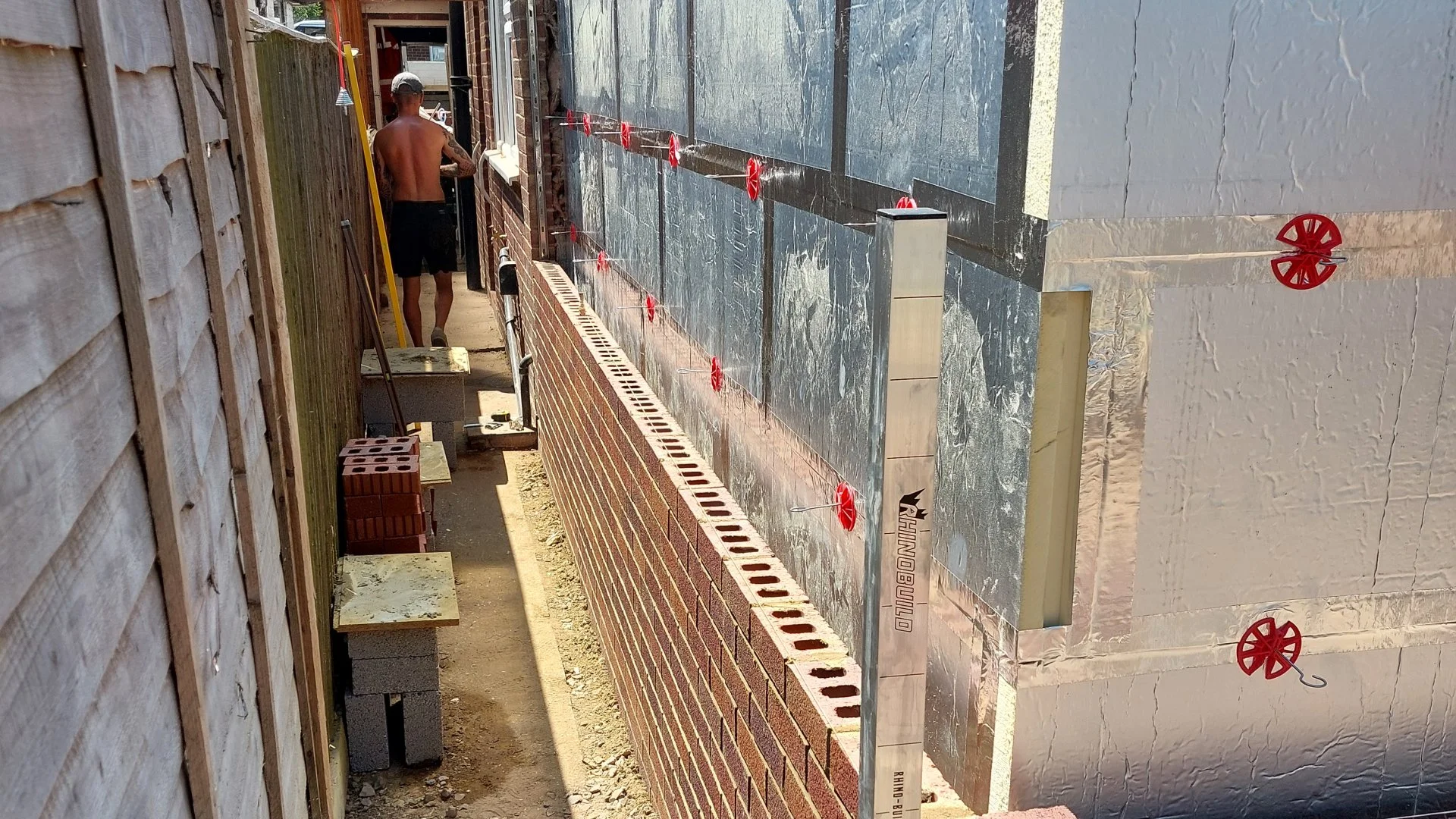 A construction worker working on a wall between two buildings in a narrow alleyway. The worker is shirtless and wearing a cap, with construction tools, bricks, and insulation materials visible.
