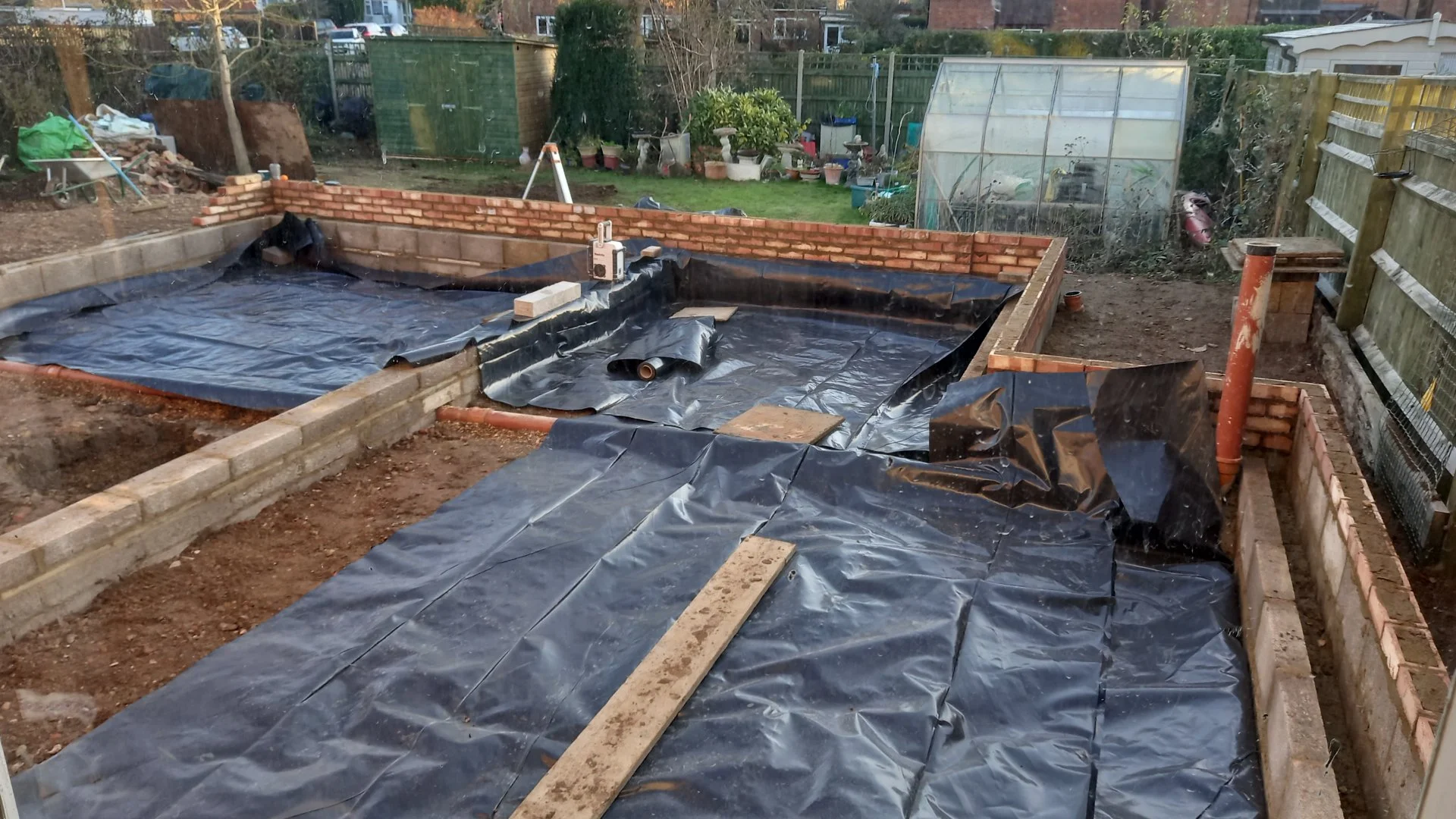 A backyard construction site with a partially built brick wall surrounding a sunken area, covered with black waterproofing membrane, and wooden planks and bricks scattered around. Garden and storage sheds are visible in the background.