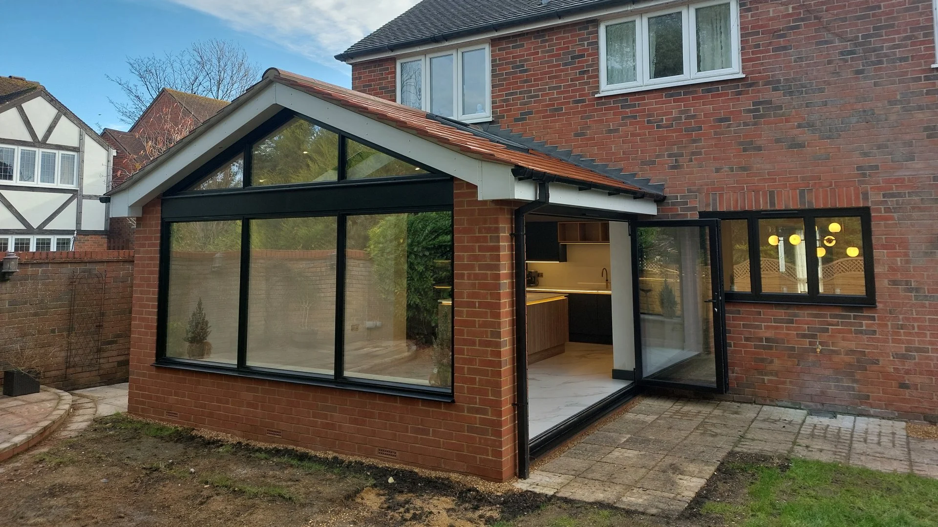 Backyard view of a brick house with a modern glass extension. The extension features large glass windows and a sliding glass door leading to the patio. The house has white-framed windows on the upper floor, and the backyard has a small patch of dirt 