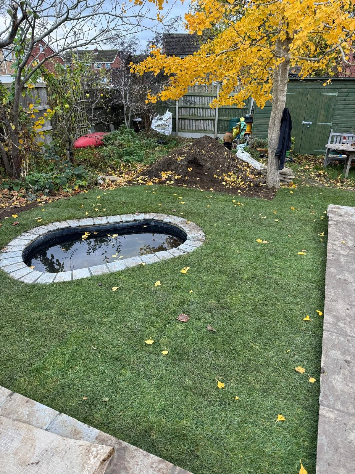 Backyard with a small pond surrounded by artificial grass, scattered autumn leaves, a tree with yellow leaves, and a garden bed with a pile of soil and gardening supplies.