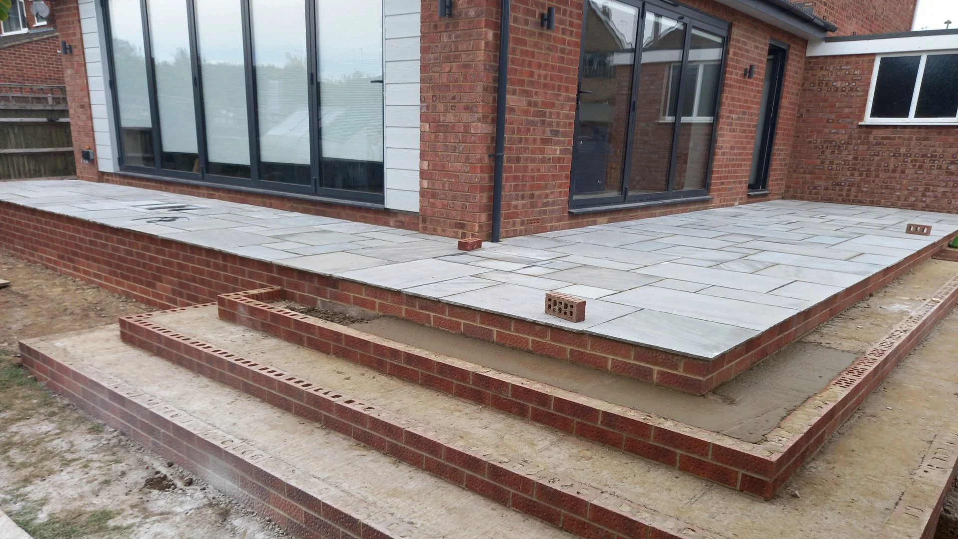 Construction site with a newly built brick patio outside a house with large sliding glass doors and windows.