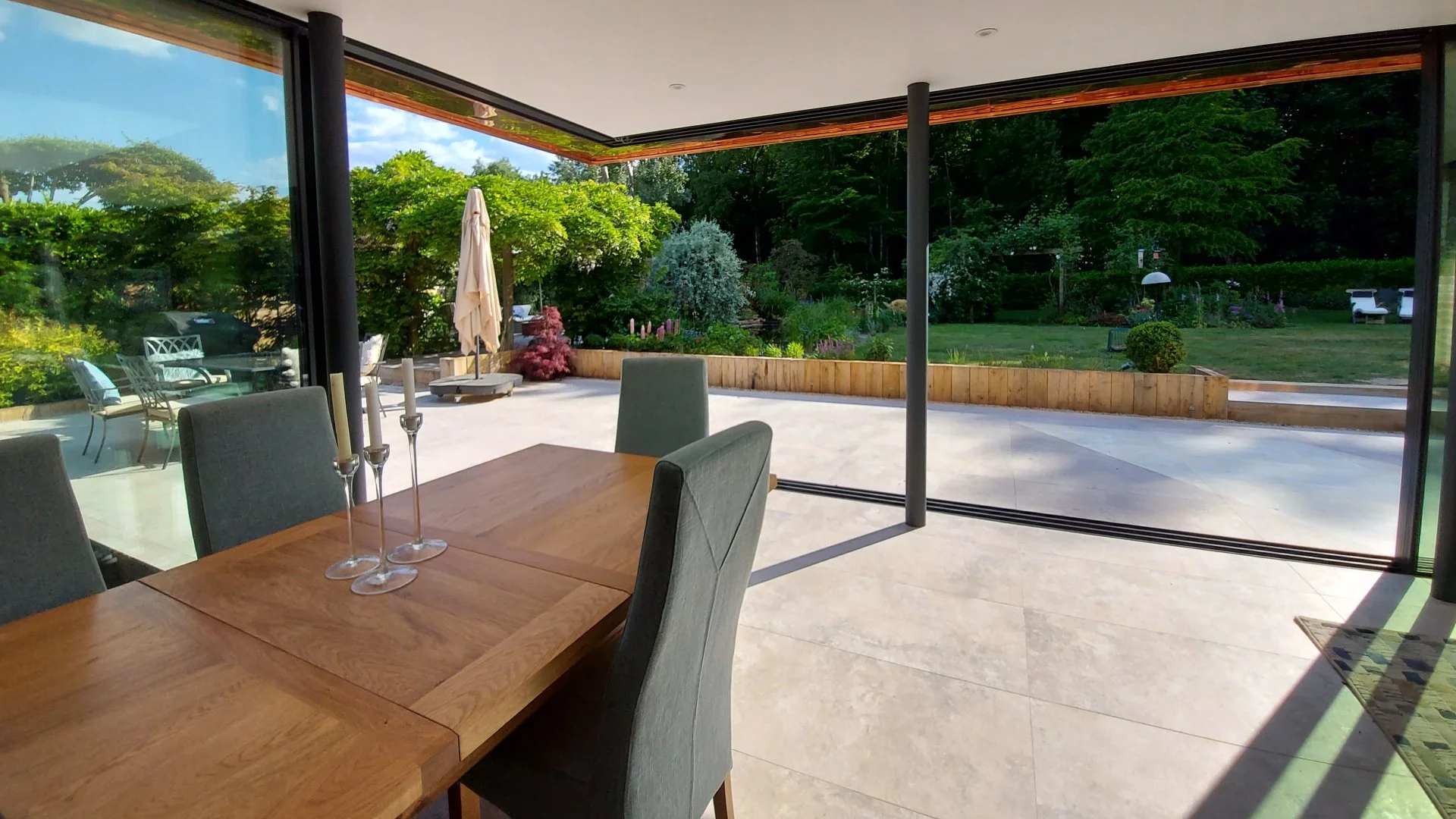A view from inside a modern house, looking through large glass doors at a backyard with patio furniture, a garden with plants and trees, and a grassy lawn.