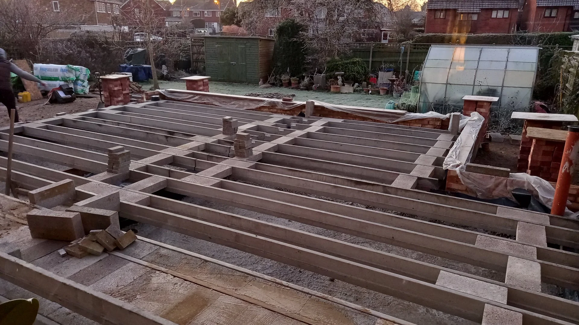 Backyard construction site with wooden and concrete framework for a deck or patio, surrounded by garden tools, plants, and a greenhouse.