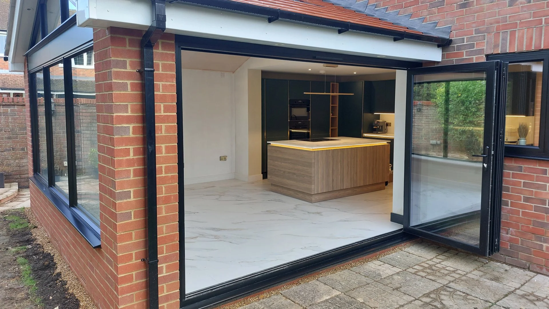 Modern kitchen with a central island, dark cabinets, built-in oven, coffee machine, and glass doors opening to an outdoor patio. White marble floor with slight veining.