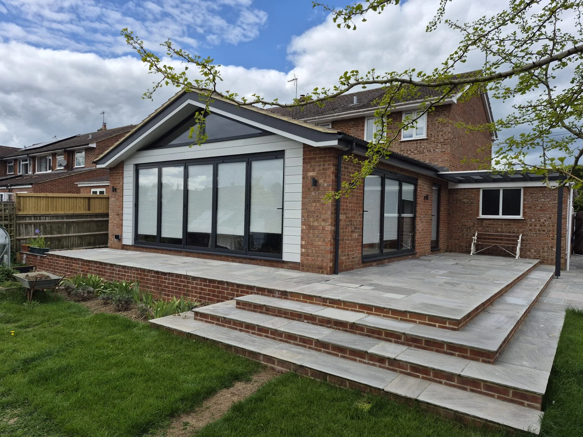 Backyard patio with brick and white siding house, large glass sliding door, tiled steps, green lawn, garden bed, and a wooden bench, under a cloudy sky.