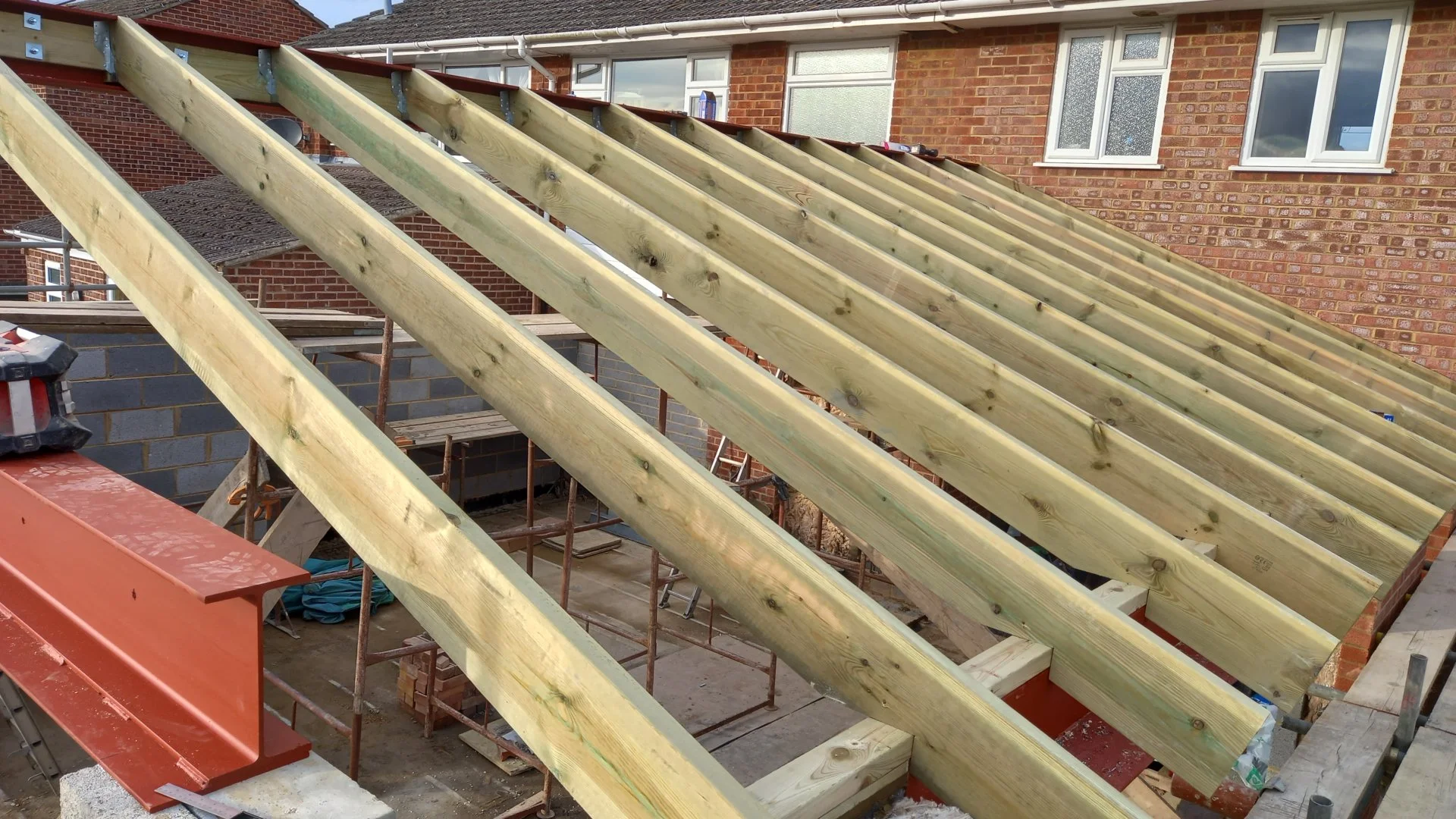 Construction site with wooden roof rafters being installed on a residential building. The house has red brick exterior walls and multiple windows. Scaffolding surrounds the building.